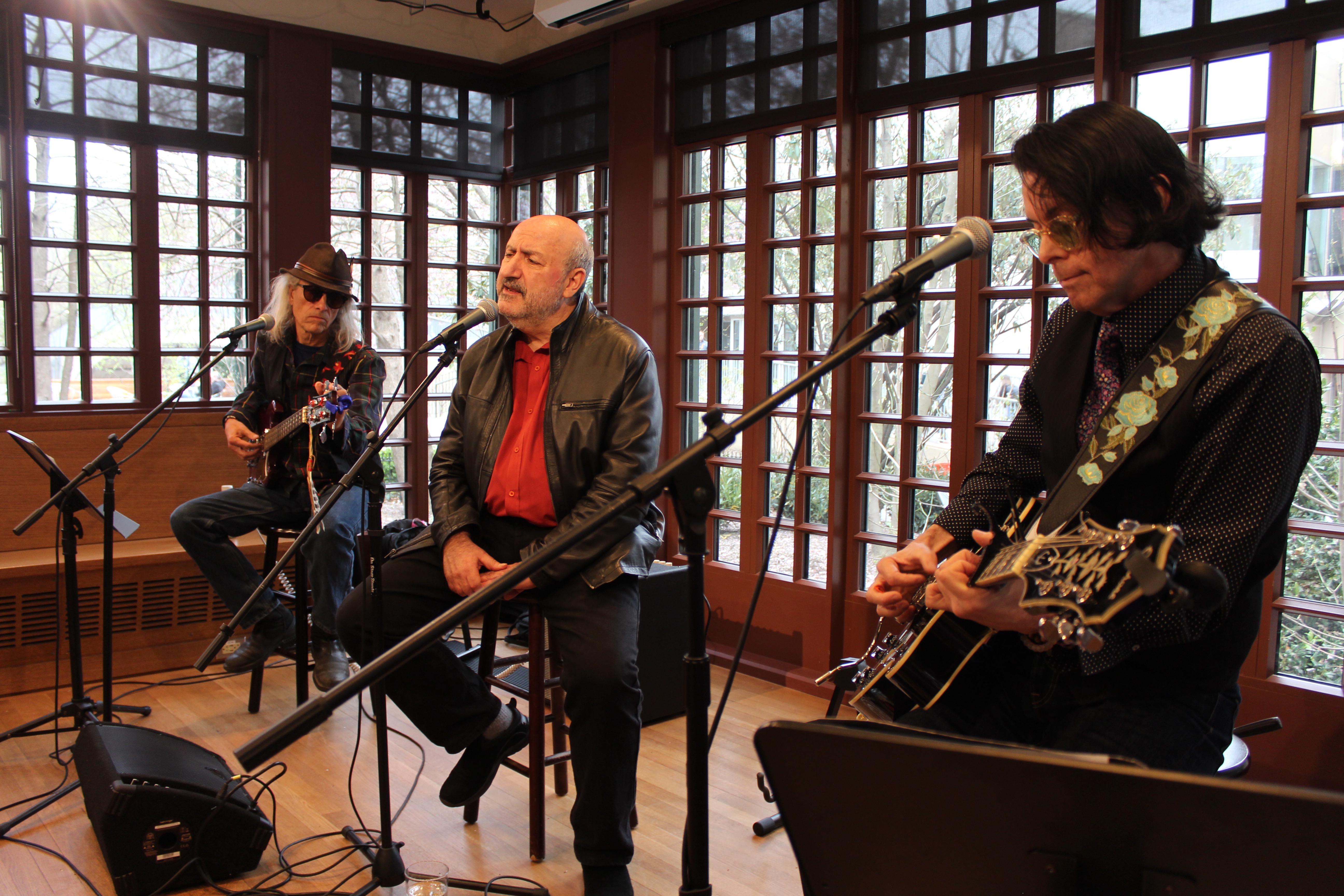 Three people sitting on high stools on a stage surrounded by windows, two playing guitars and one speaking or singing into a microphone. 