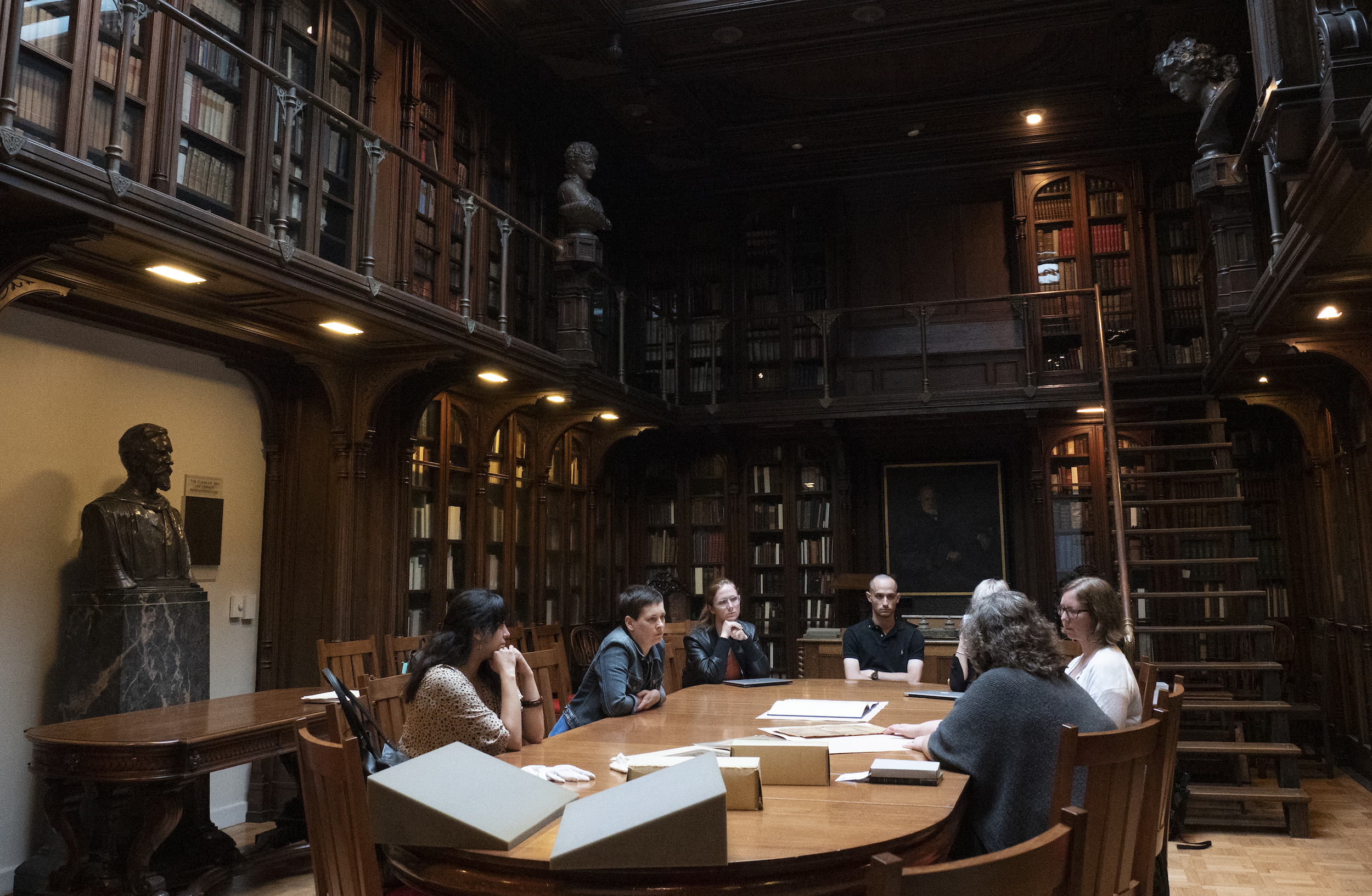 A group of people sitting around a rectangular wooden table on the bottom floor of a two-story room in a library adorned with books and busts.