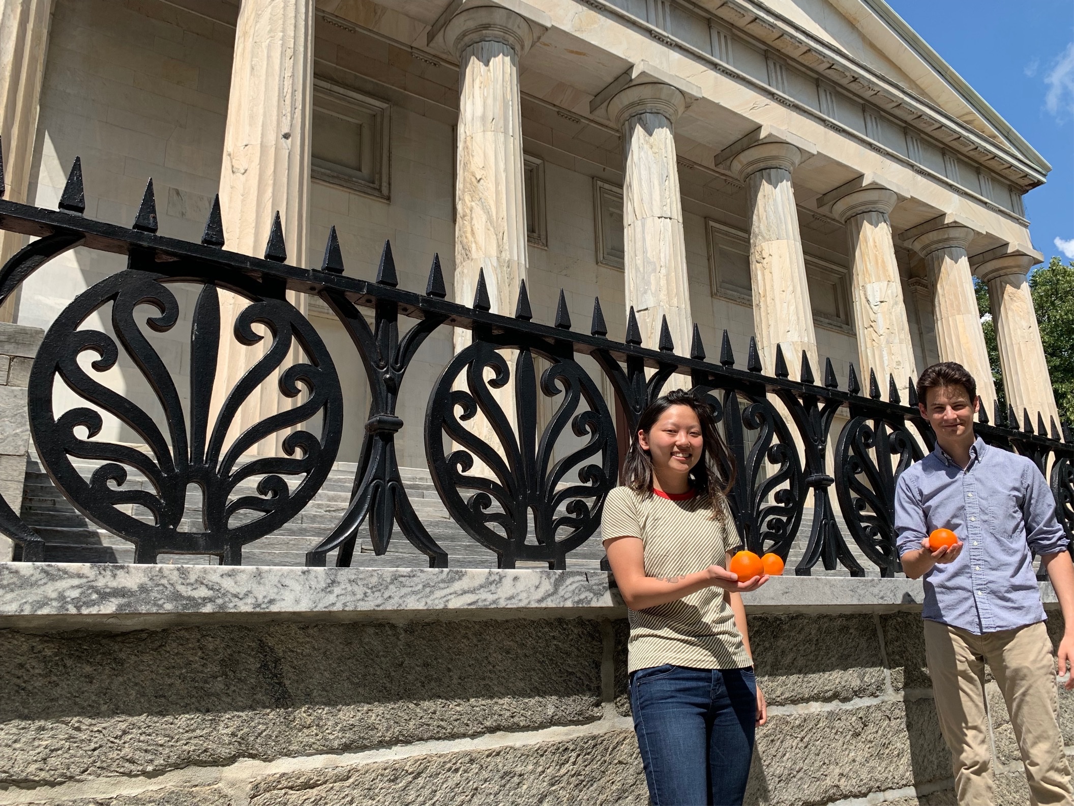 Two students holding oranges standing in front of historic building with stone columns.
