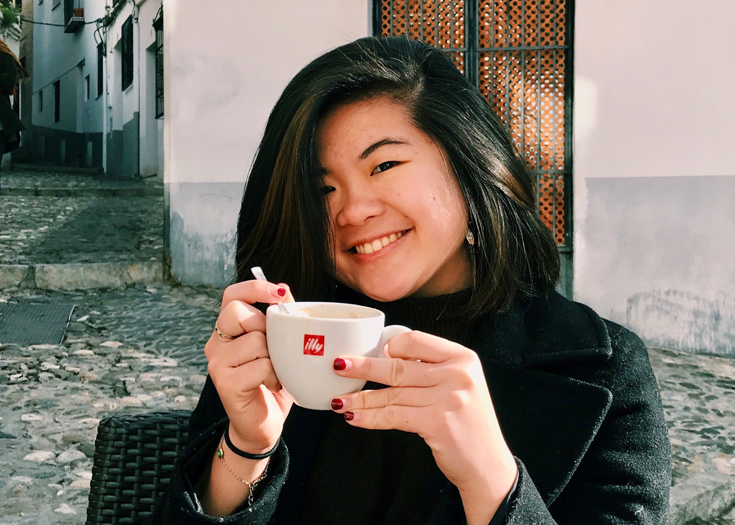 Student sitting outside holding a coffee cup and smiling. 