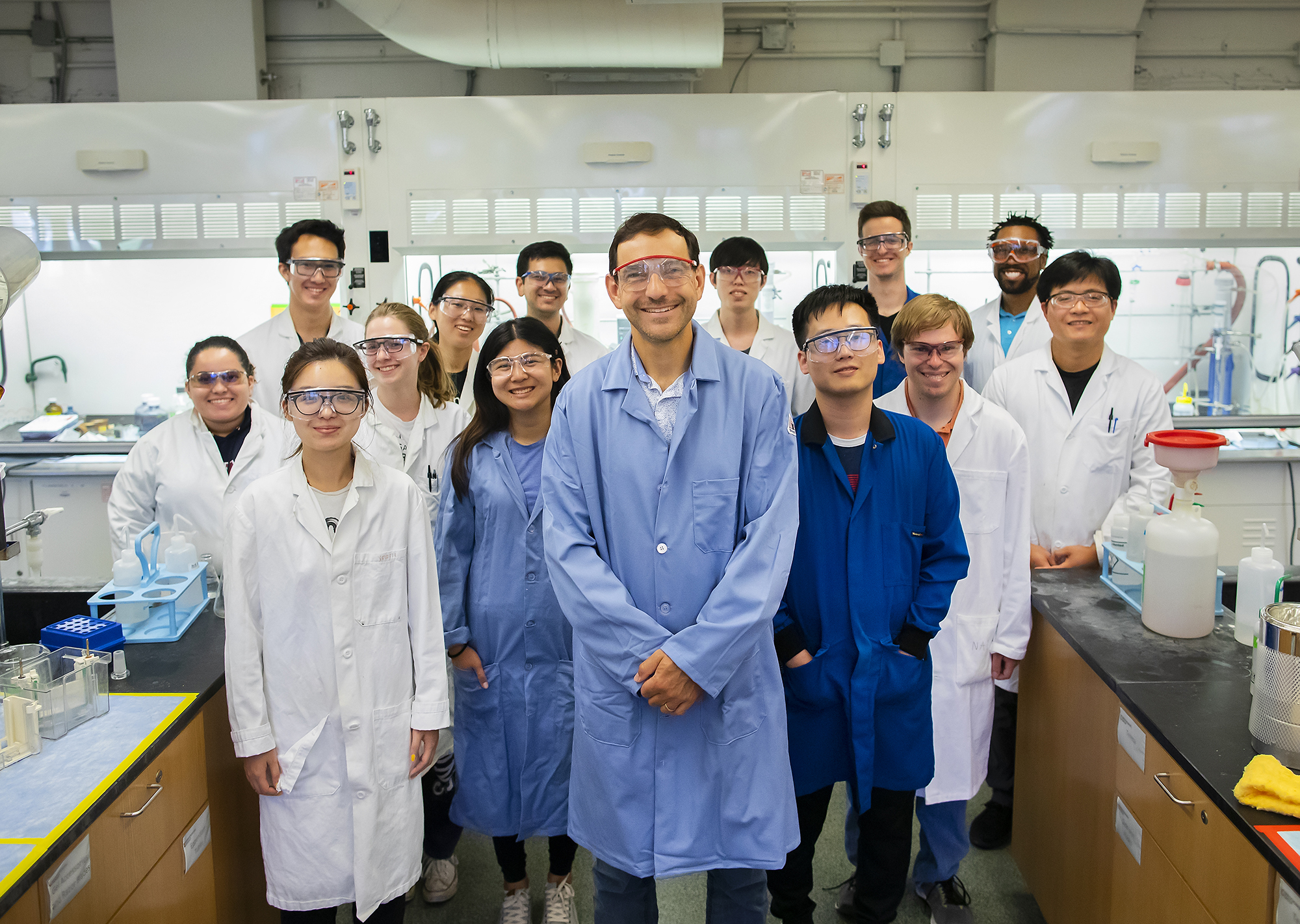 a group of people standing in a chemistry lab wearing lab coats and goggles