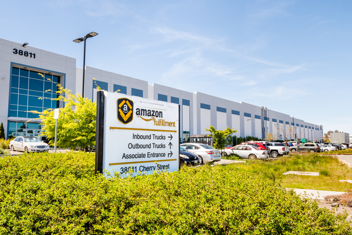 View of an Amazon fulfillment center outside in the sun with a sign and parking lot out front.