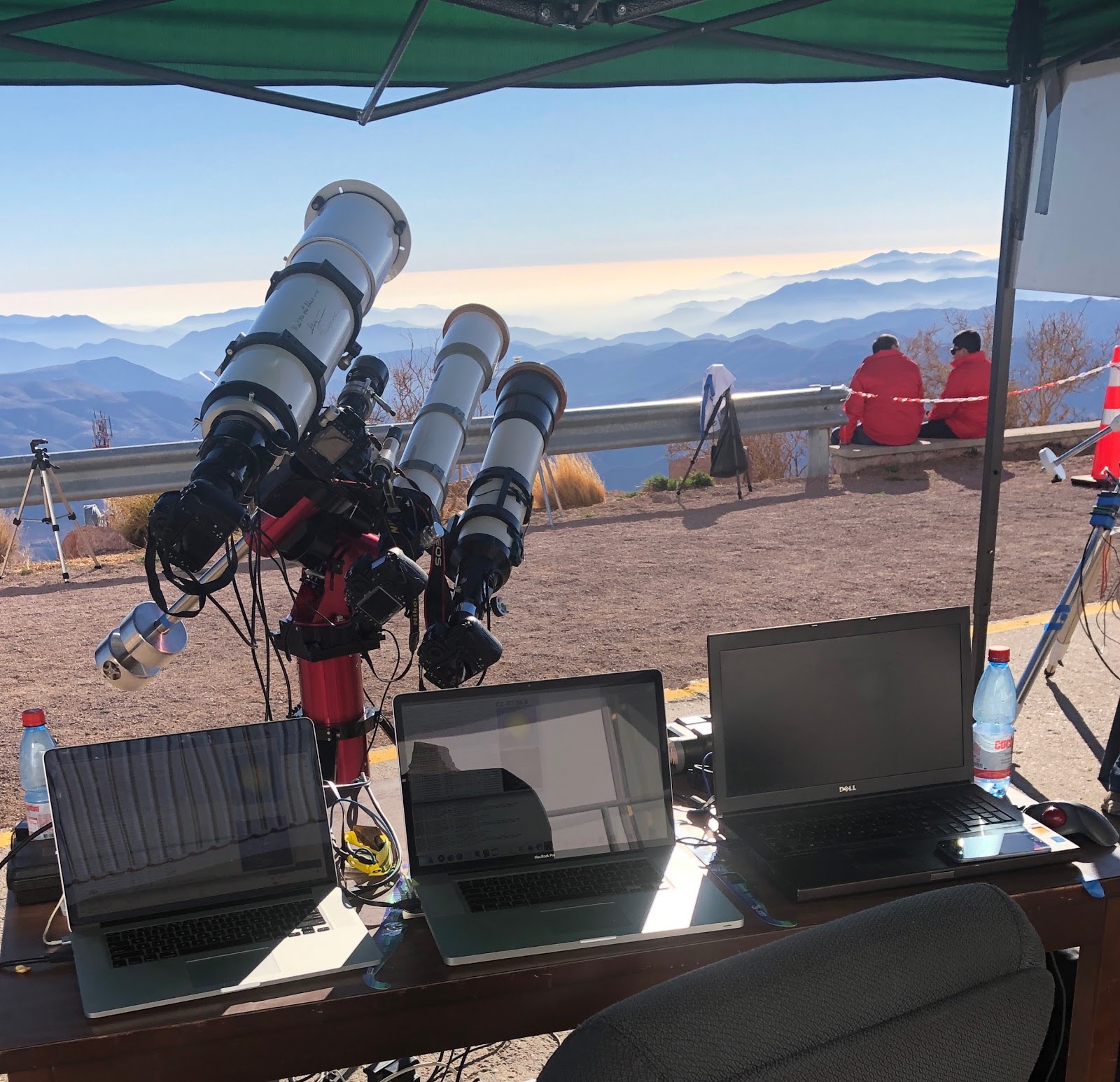 three telescopes positioned above the horizon of mountains with three laptops in the foreground