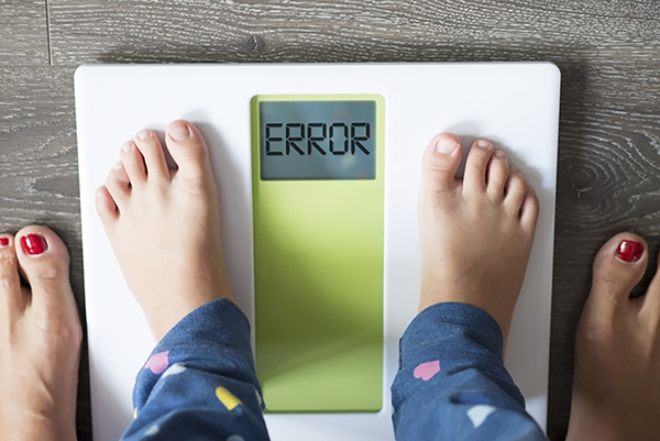 small child stands on scale with error message with adult's feet alongside the scale, symbolizing early weight stigma