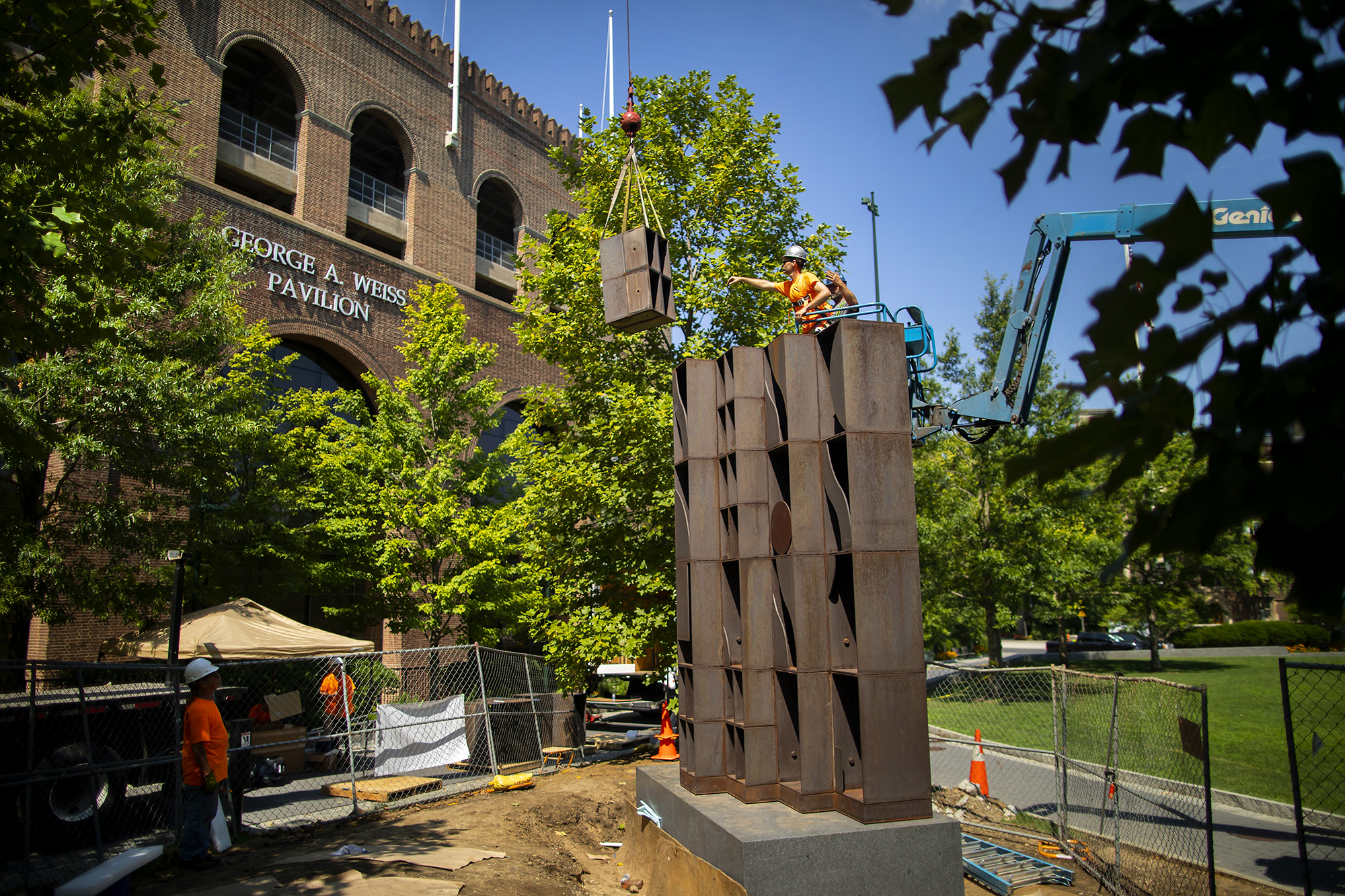 Sculpture made of metal cubes on a base with a construction worker on top reaching for another cube suspended by a crane. 