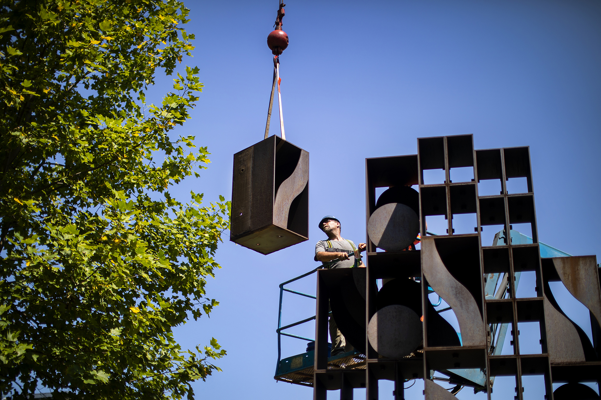 Large cube of metal suspended in the air by a crane as a construction worker looks at it from the top of a sculpture made of similar metal cubes. 