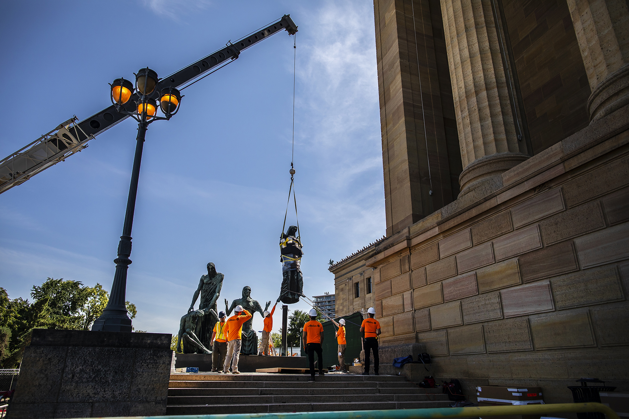 View of sculpture with three sets of figures and one on right is being lifted with a large crane. Several construction workers with hardhats are standing around it, at the top of steps next to a large stone building with columns.