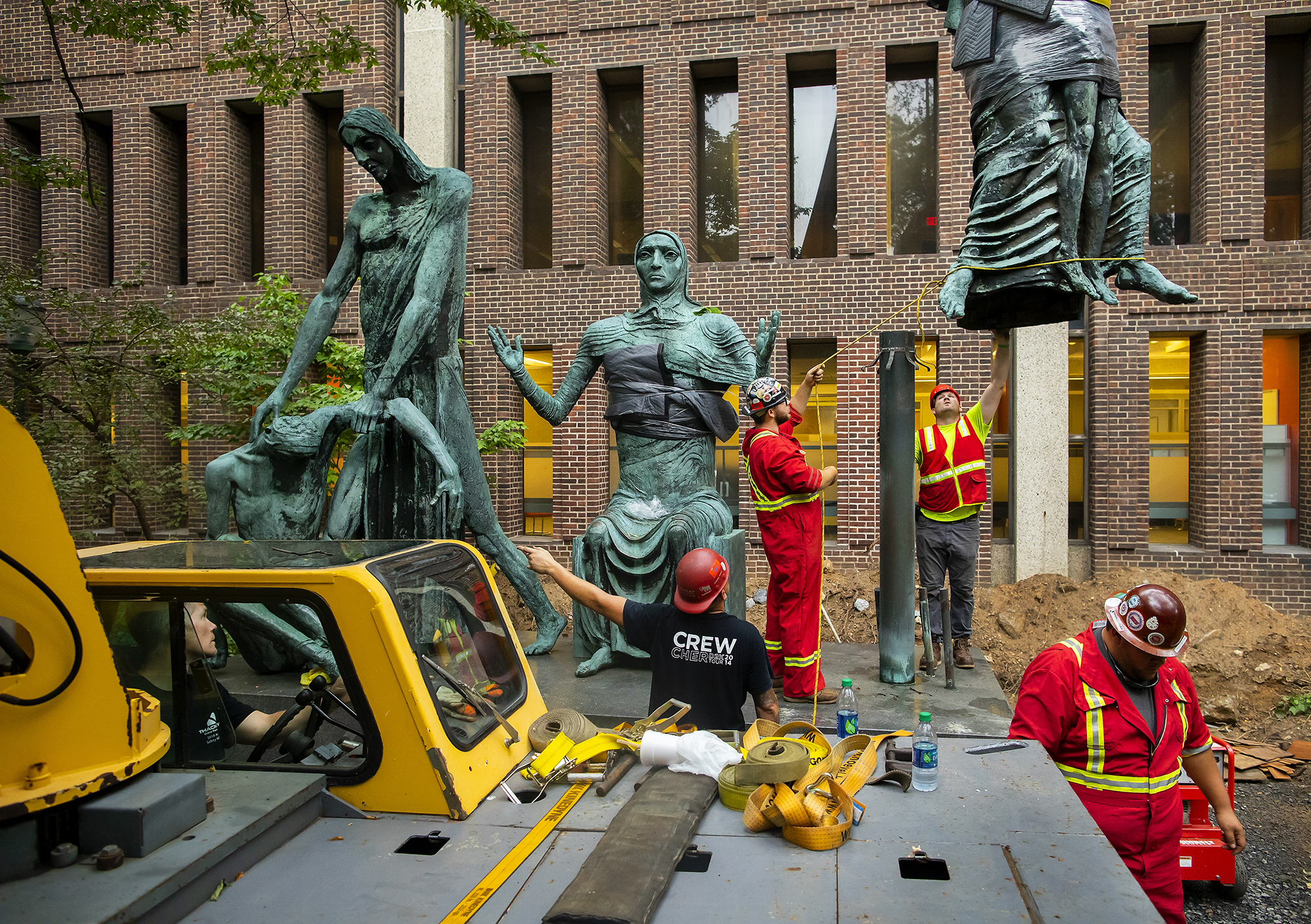 A large sculpture with two figures in place on the left, one in the middle, and two more being lowered into place with two construction workers reaching up toward the dangling feet, and other workers with the cab of a large crane.