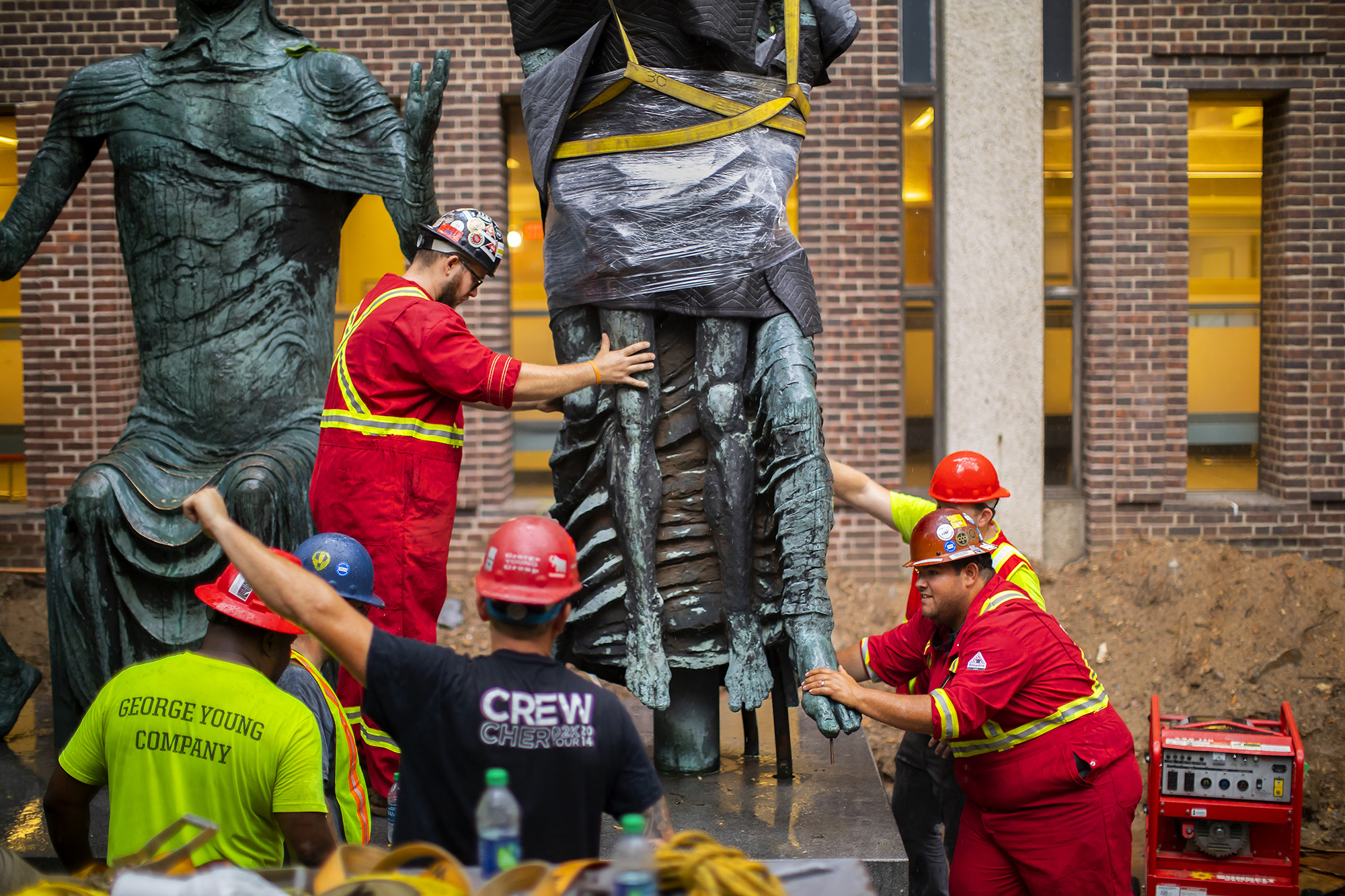 A large sculpture figure is lowered onto a support rod by several construction workers. 
