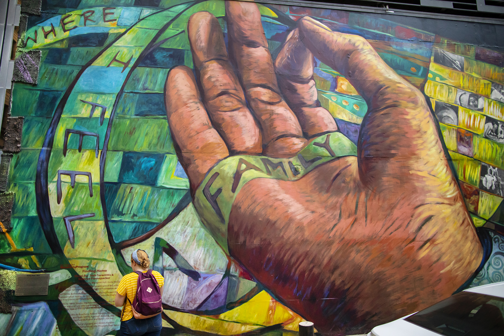 One student standing in front of large mural depicting a hand with a ribbon of color that reads where I feel family. 