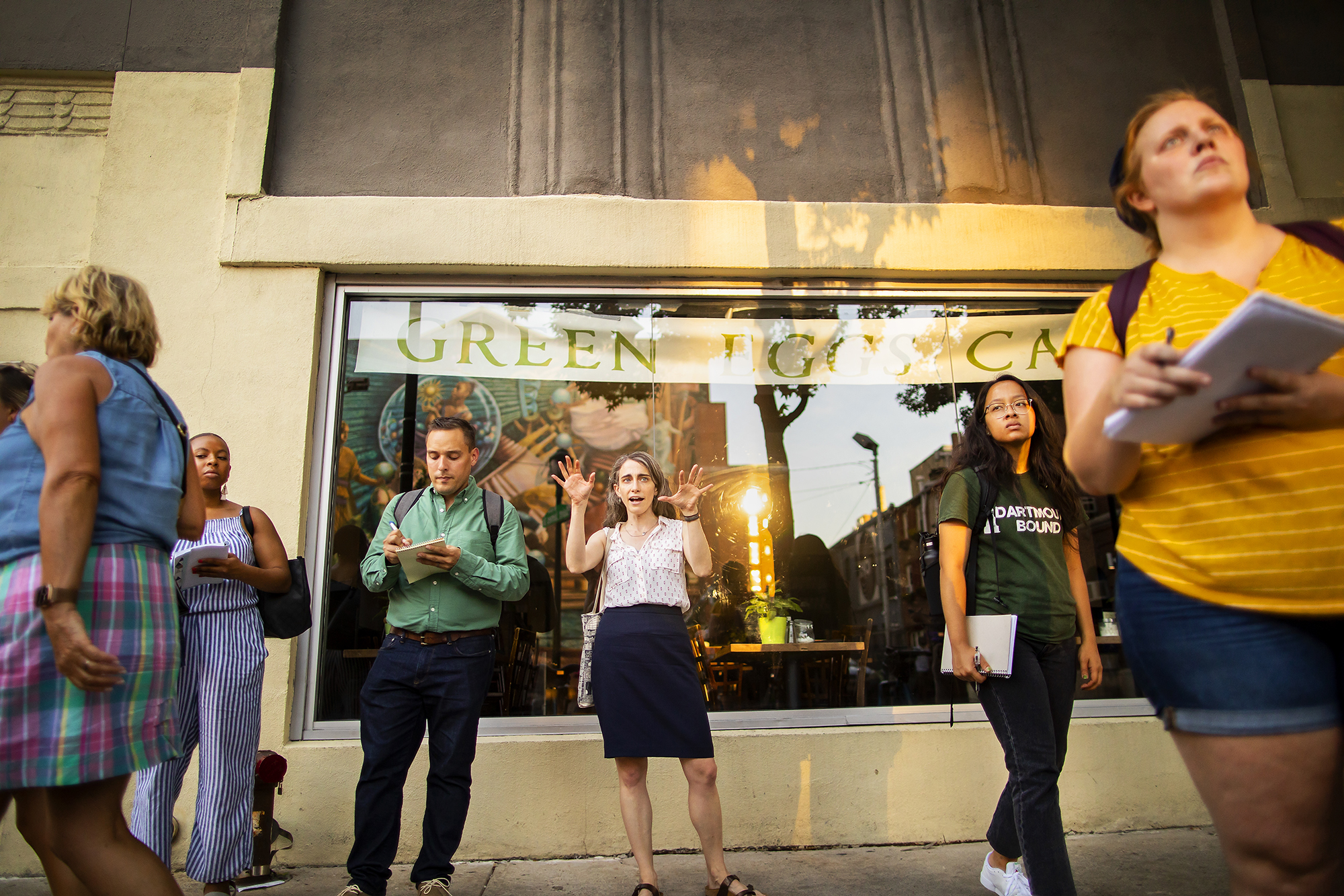 Five people, four with notebooks and one speaking, standing in front of a storefront window reflecting a mural across the street.