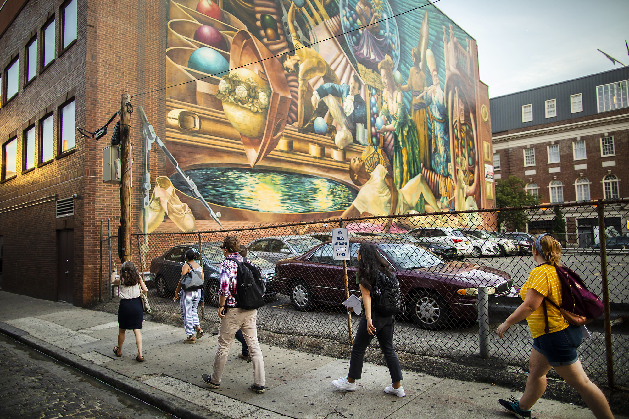 Five people walking on a sidewalk next to a parking lot looking up at a colorful mural on the wall of the adjacent building. 