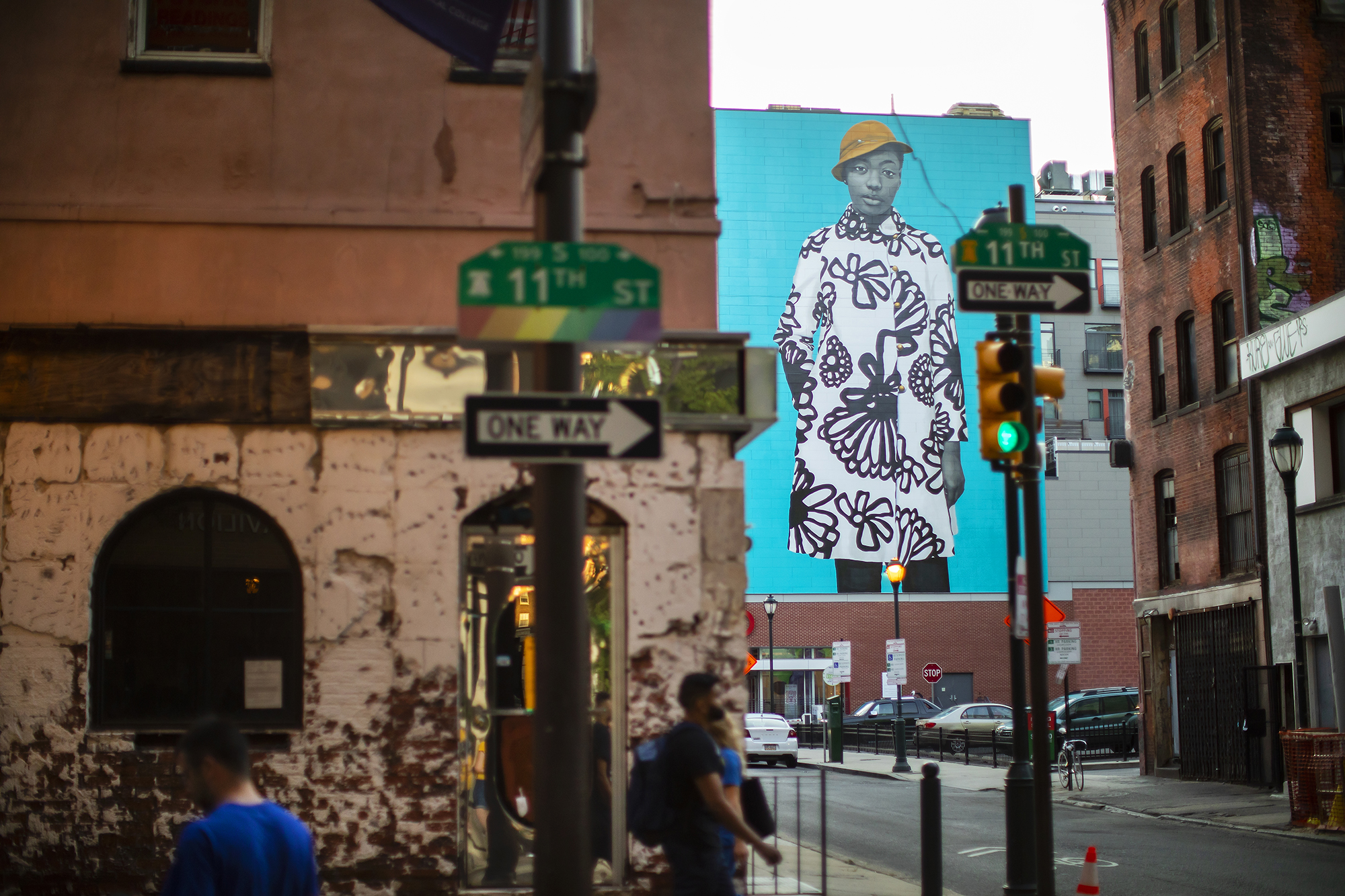 A mural featuring a young woman can be seen beyond brick buildings and a traffic light and signs including 11th street and one way.