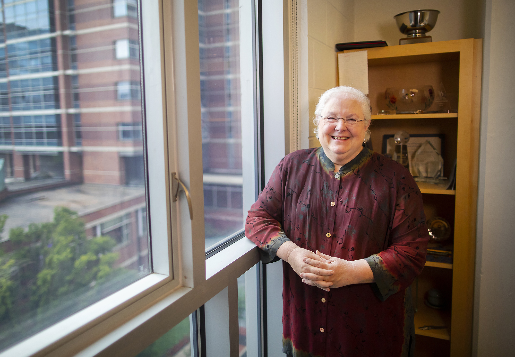 Person standing at a large window with a bookshelf behind. ON the bookshelf are several awards and plaques.