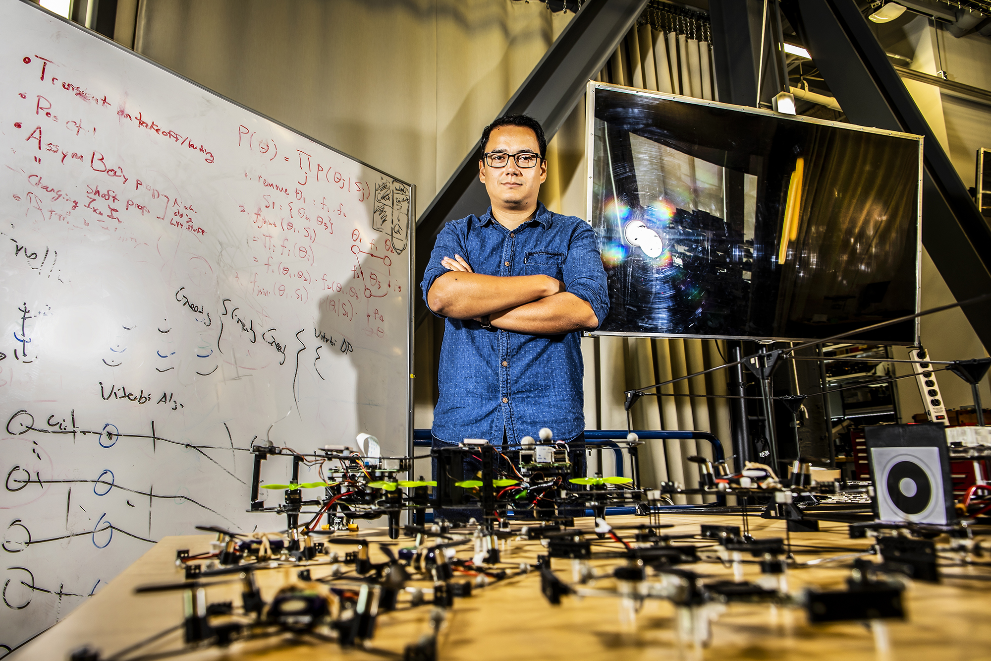 david saldana behind a table of his modular flying robots in front of a white board of equations and a TV screen