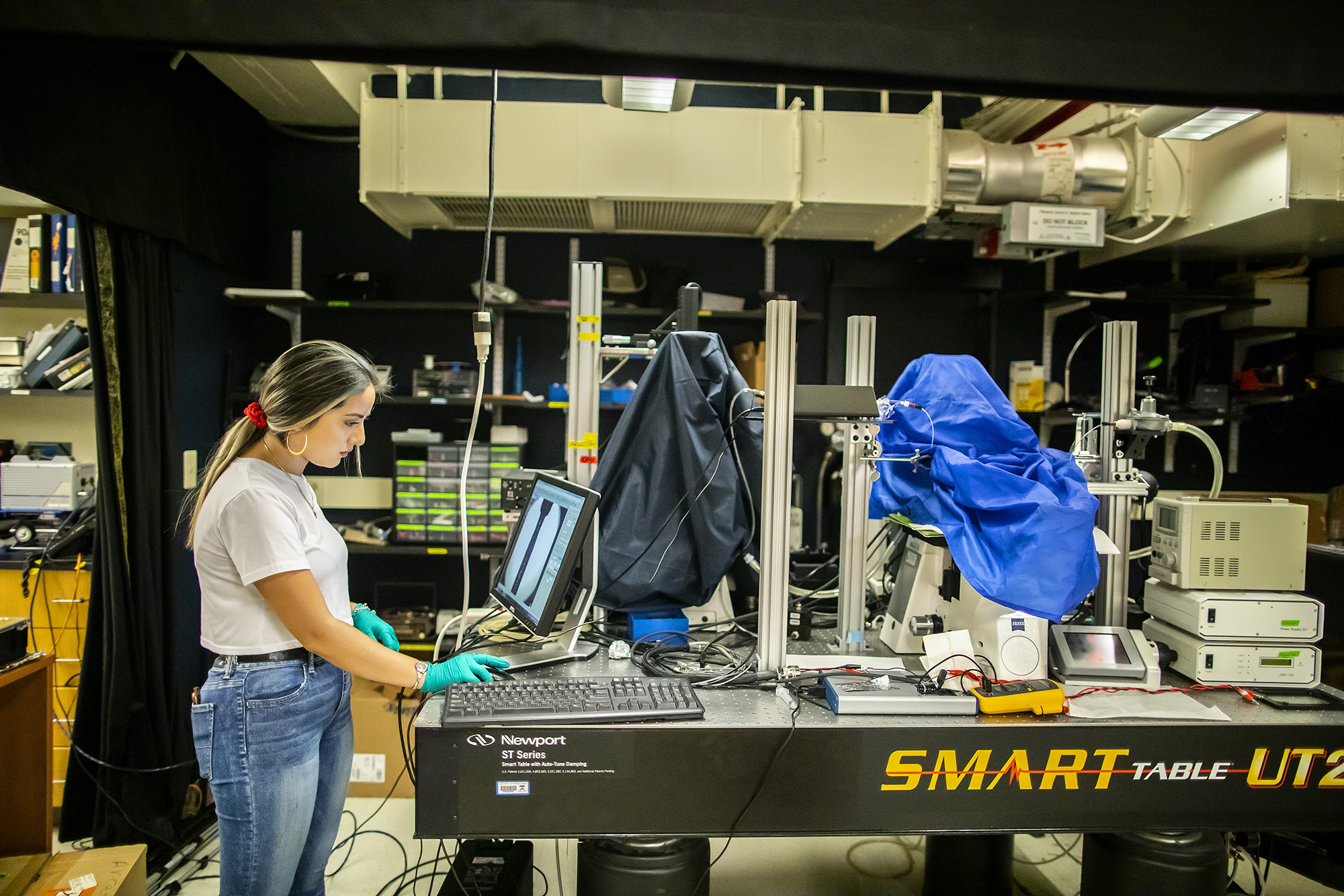 angelica padilla working in a lab on a crowded optics table looking at a computer