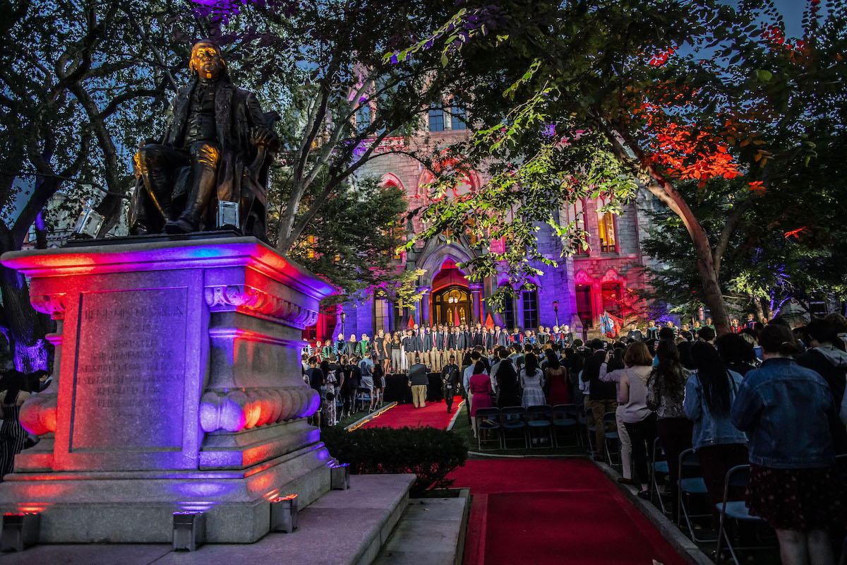 View of the Ben Franklin statue and College Hall lit in red and purple lights at dusk during convocation.
