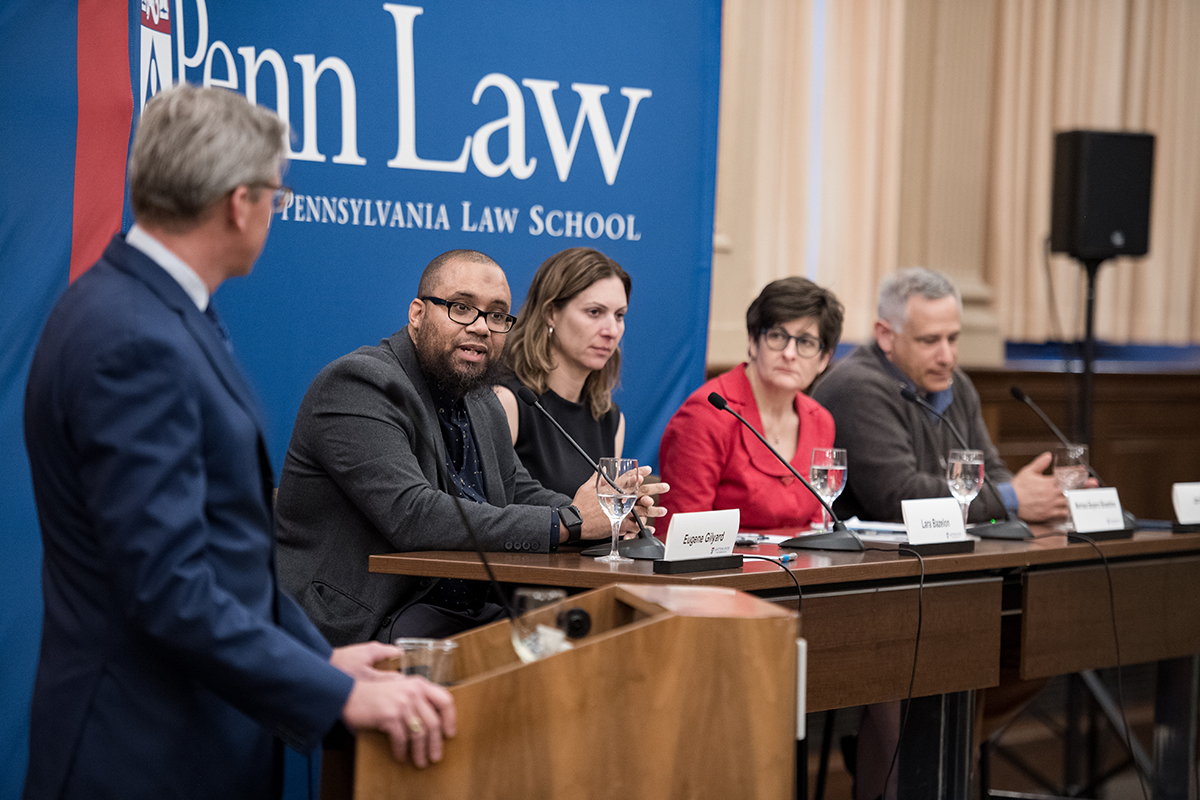 Panel conference with John Hollway at podium, seated to his left are Eugene Gilyard, speaking, and three other panel members, in front of a Penn Law banner.