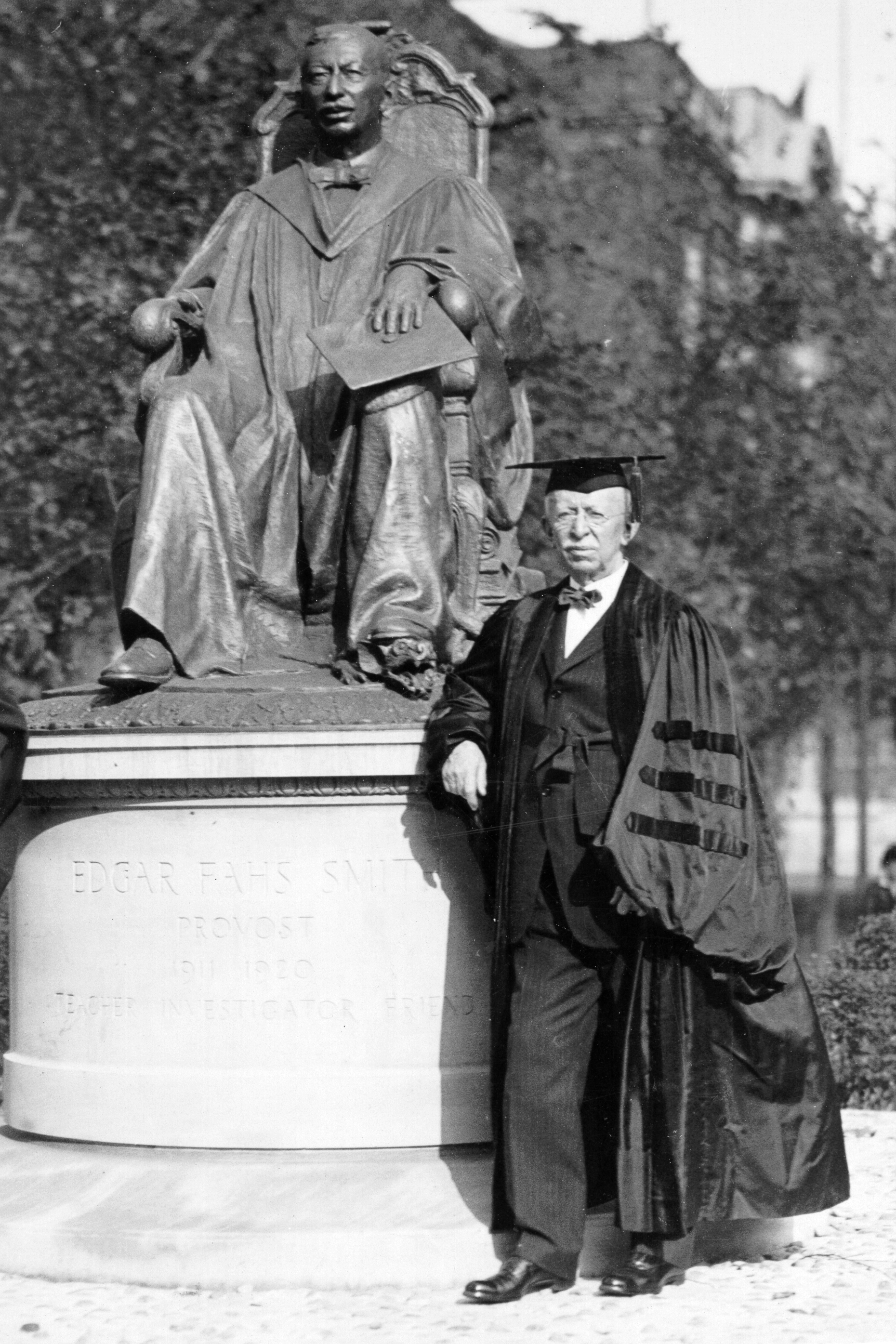 edgar fahs smith posing in front of a statue of himself, he is wearing a cap and gown and stands at the level of the statue's feet