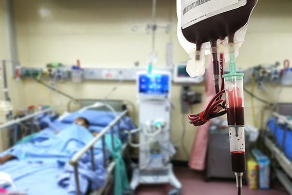 A blood bag hangs in the foreground, as a transfusion for a hospital patient lying in bed in the background.