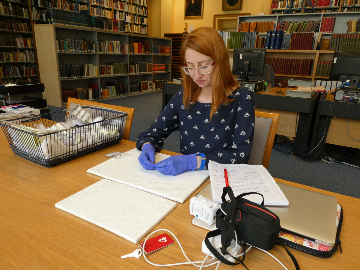 In a room with stacks of books on bookshelves, a person sits at a table wearing rubber gloves to analyze materials sitting in a basket on her right. She holds them over two white papers, and to her left are a notepad, phone, and computer.