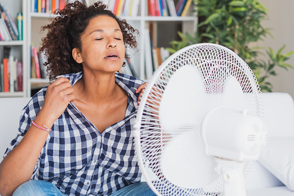 A person sits in front of a fan holding shirt front open to cool off, indicating rising temperatures