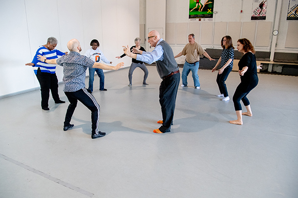 A group of people with Alzheimer's and an instructor stand in a circle in a large dance studio with their arms outstretched, bending their knees.