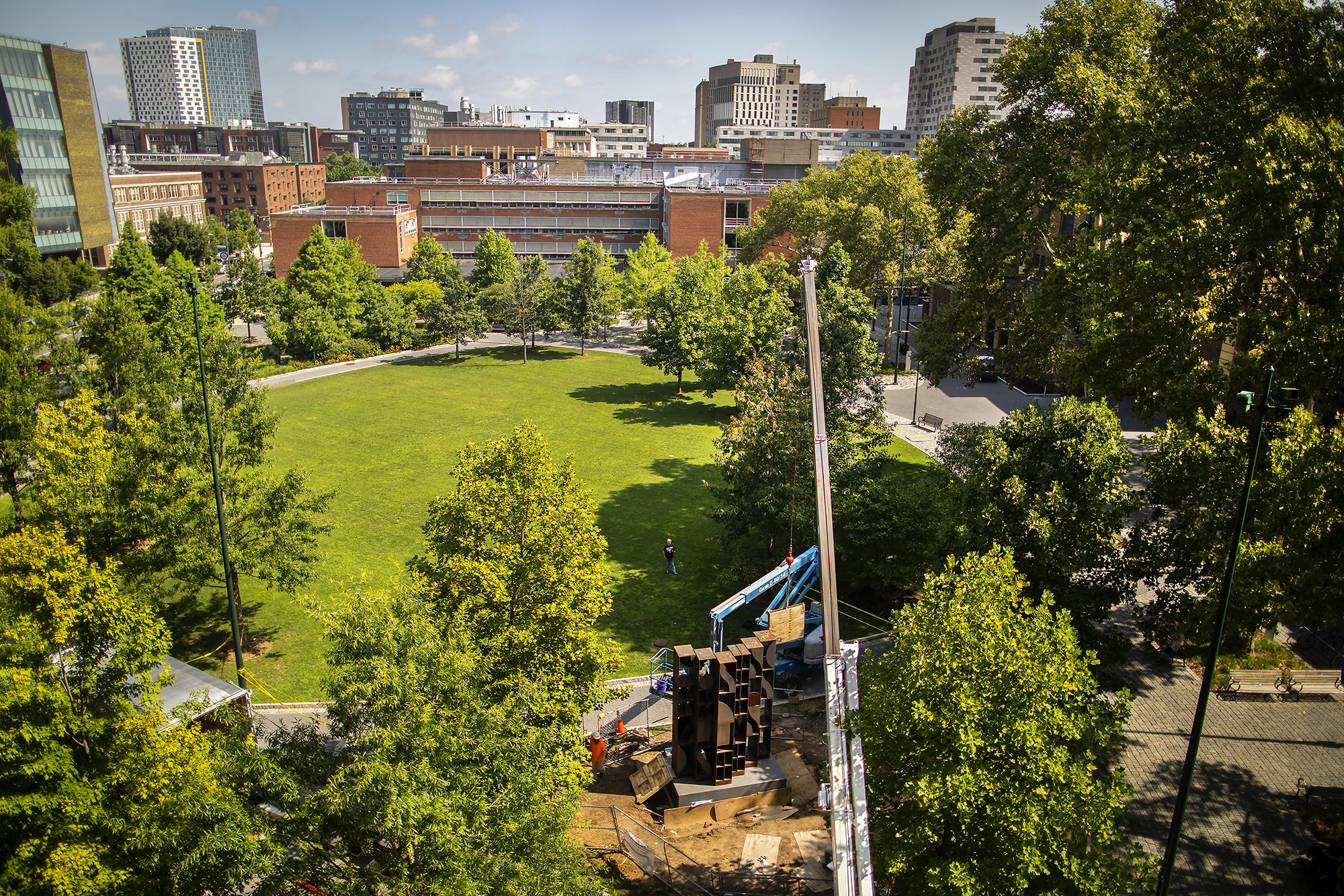 An aerial view of a large green space surrounded by trees and a sculpture under construction with the arm of a large crane above it. 