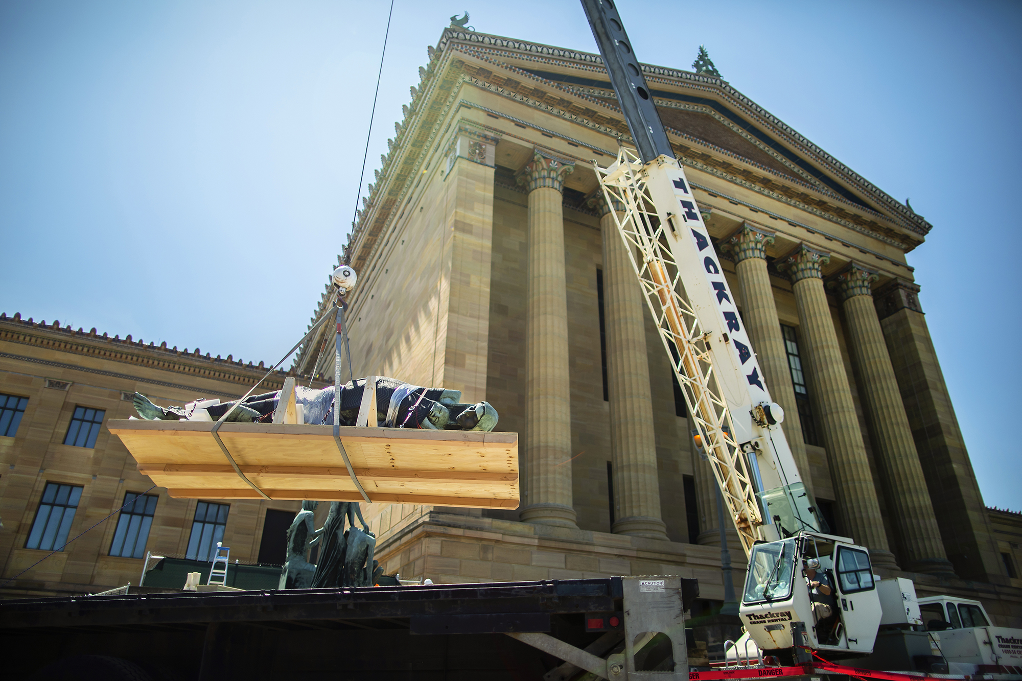 Sculpture figure strapped to a board being suspended in midair as it is lowered onto a flatbed truck with a large columned building in the background. 