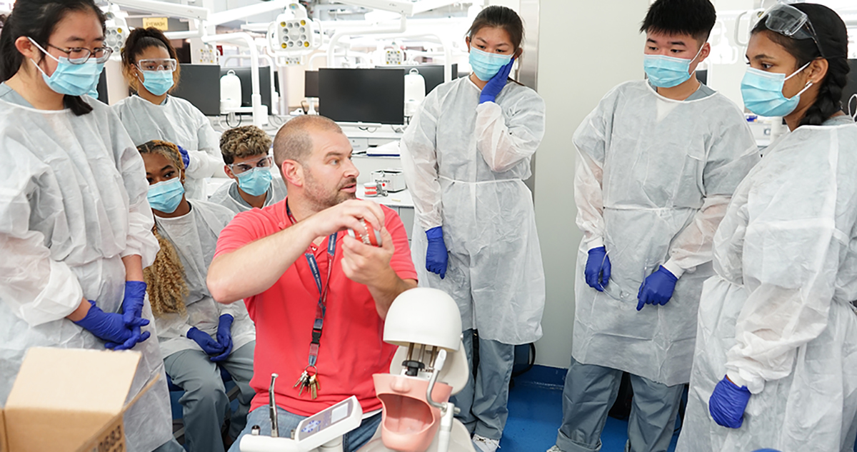 A Penn Dental Medicine instructor sits with a dental model tool in hand beside a replica of head with an open mouth while seven high school students in white lab coats, gloves and masks watch the demonstration.