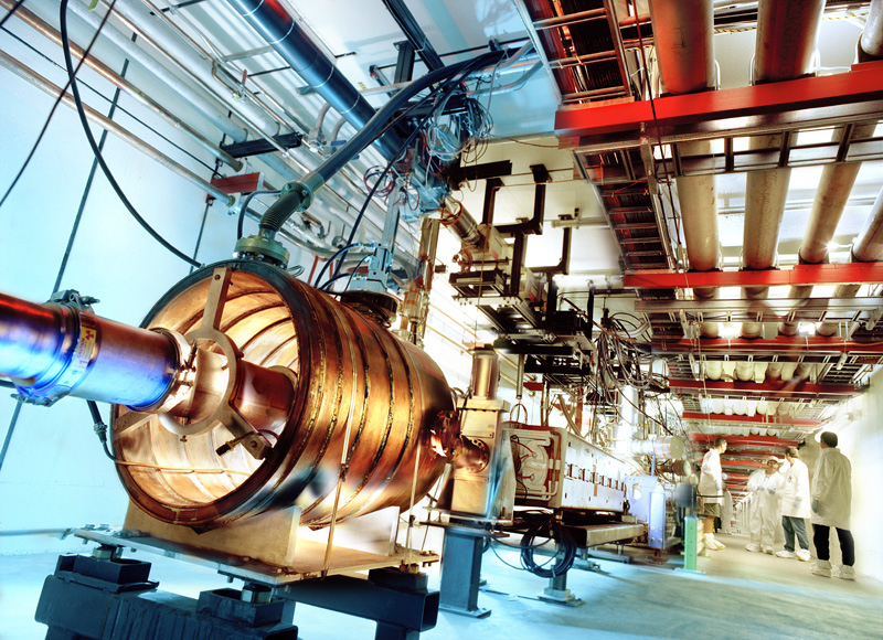 a particle accelerator, a long copper tube that follows down a basement corridor below lines of tubes and wires, a group of scientists in lab coats talk with each other on the side of the room