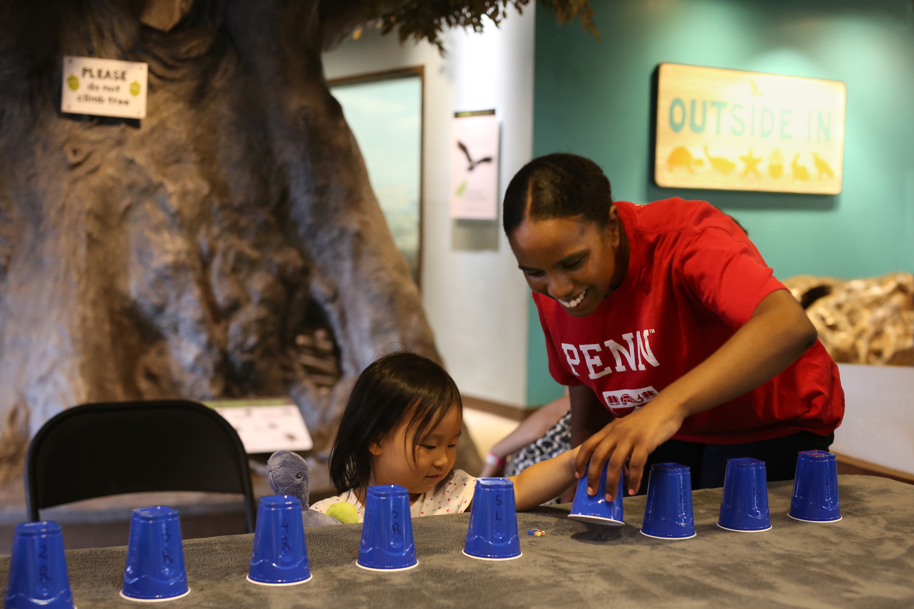 A young child at a table with an adult, playing a game with blue plastic cups. 