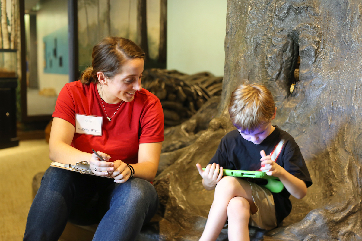 An adult and a child sit in front of a fake tree. The child plays a game on an electronic tablet as the adult watches.
