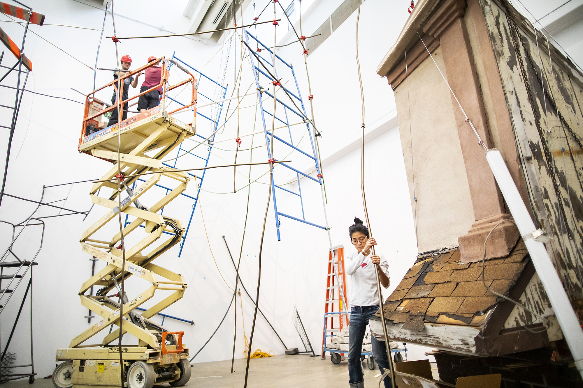 Artist with hands on long metal pipe with fragment of a building behind her and scaffolding hanging from the ceiling during sculpture installation. 