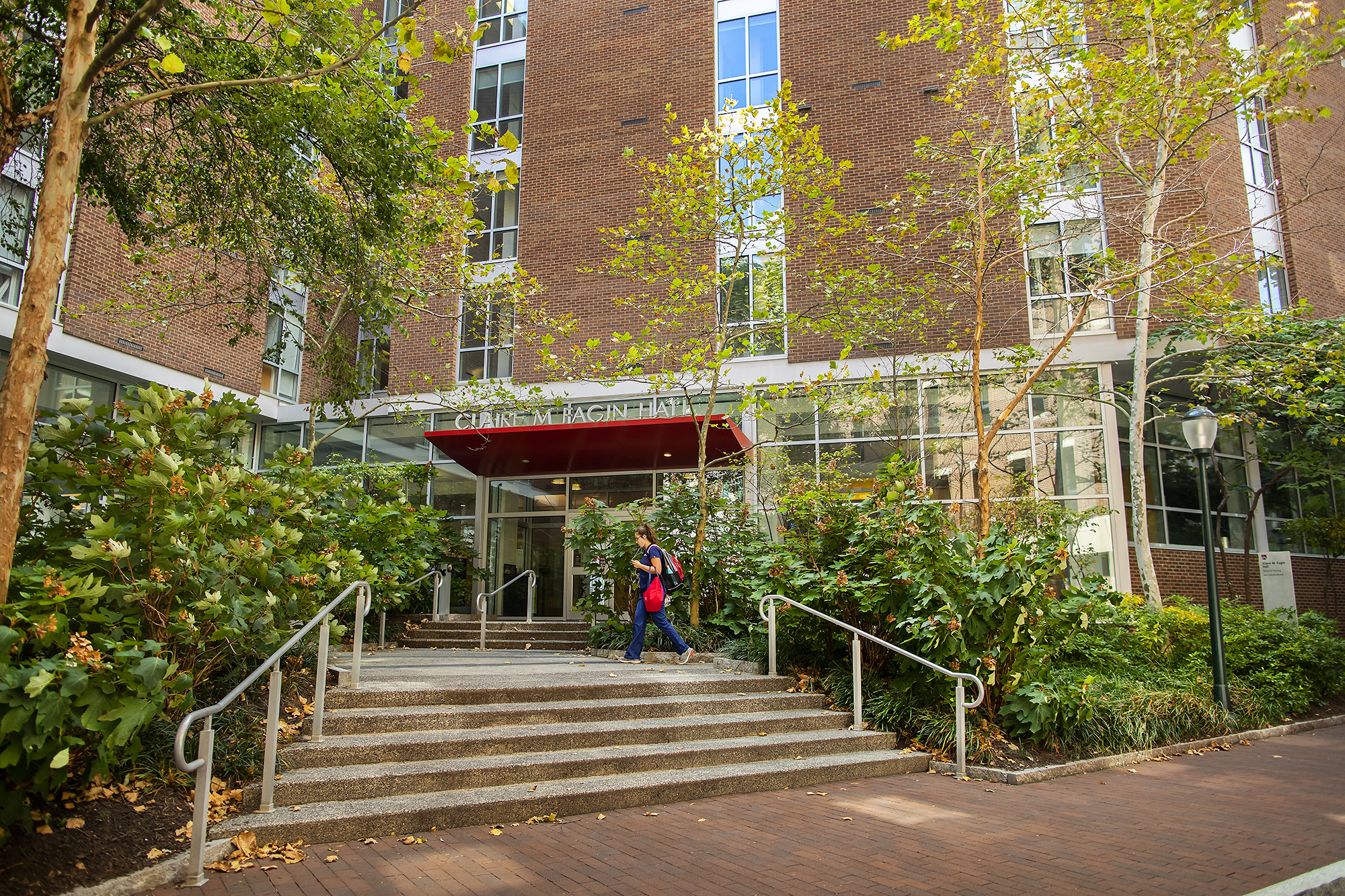 Exterior of a brick building with green trees and bushes surrounding a set of two stairs. The words "Claire M. Fagin Hall" are atop a red awning in front of the door. 
