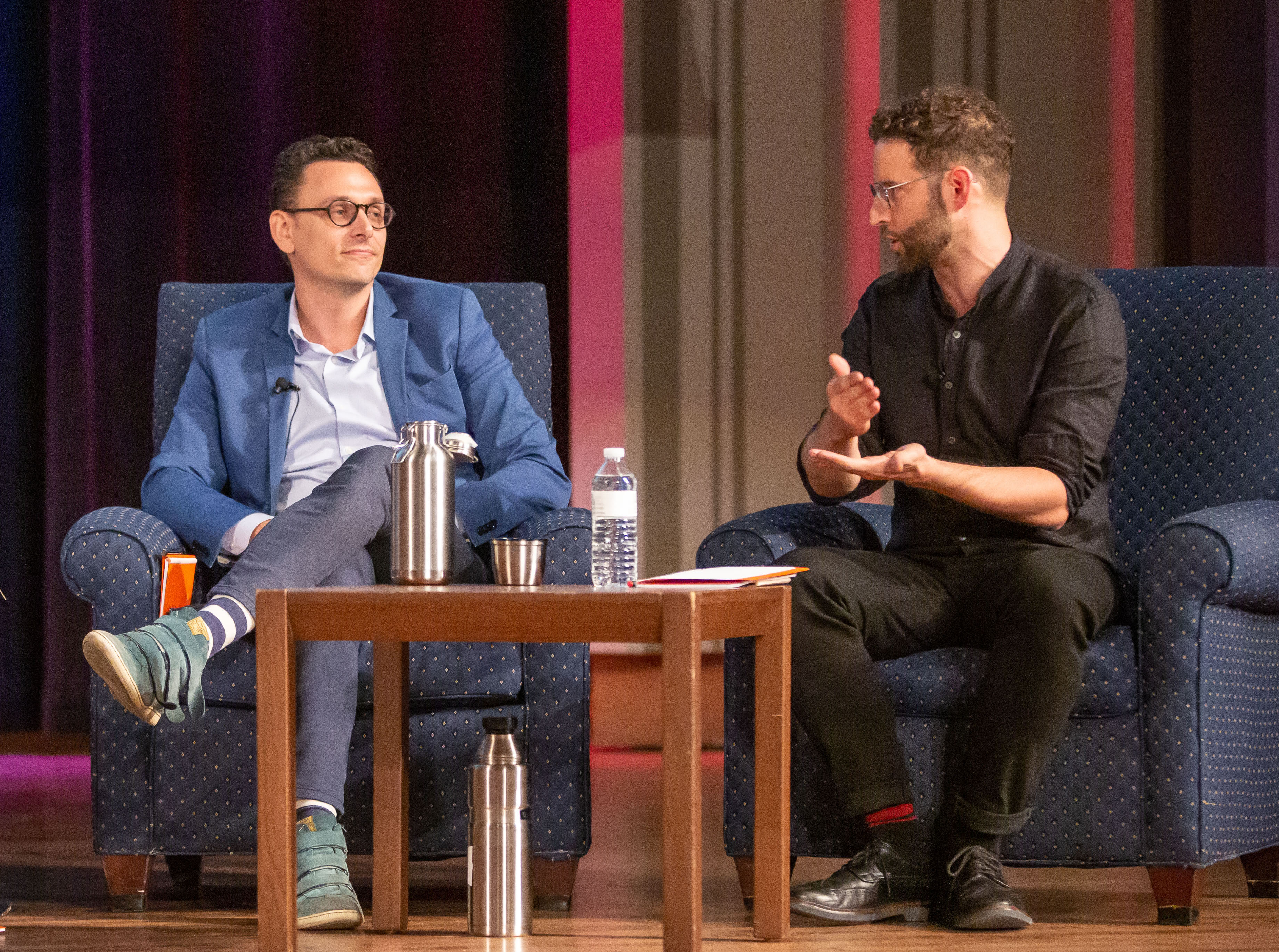 Two people sitting on a stage, one gesturing with his hands. In front of them is a brown wooden table with two water bottles.