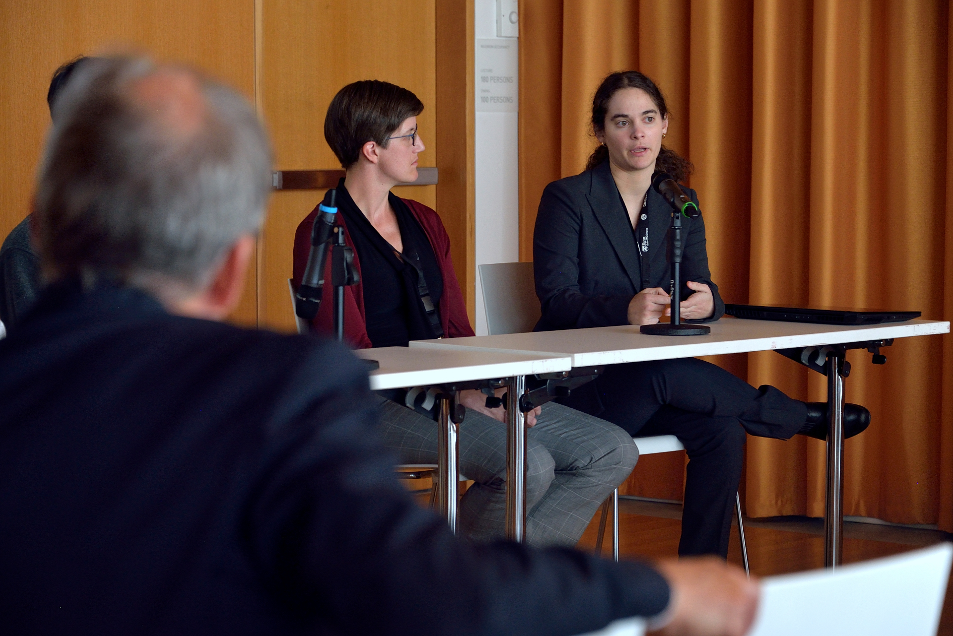 two people sit at a table in front of an audience with one talking into a microphone