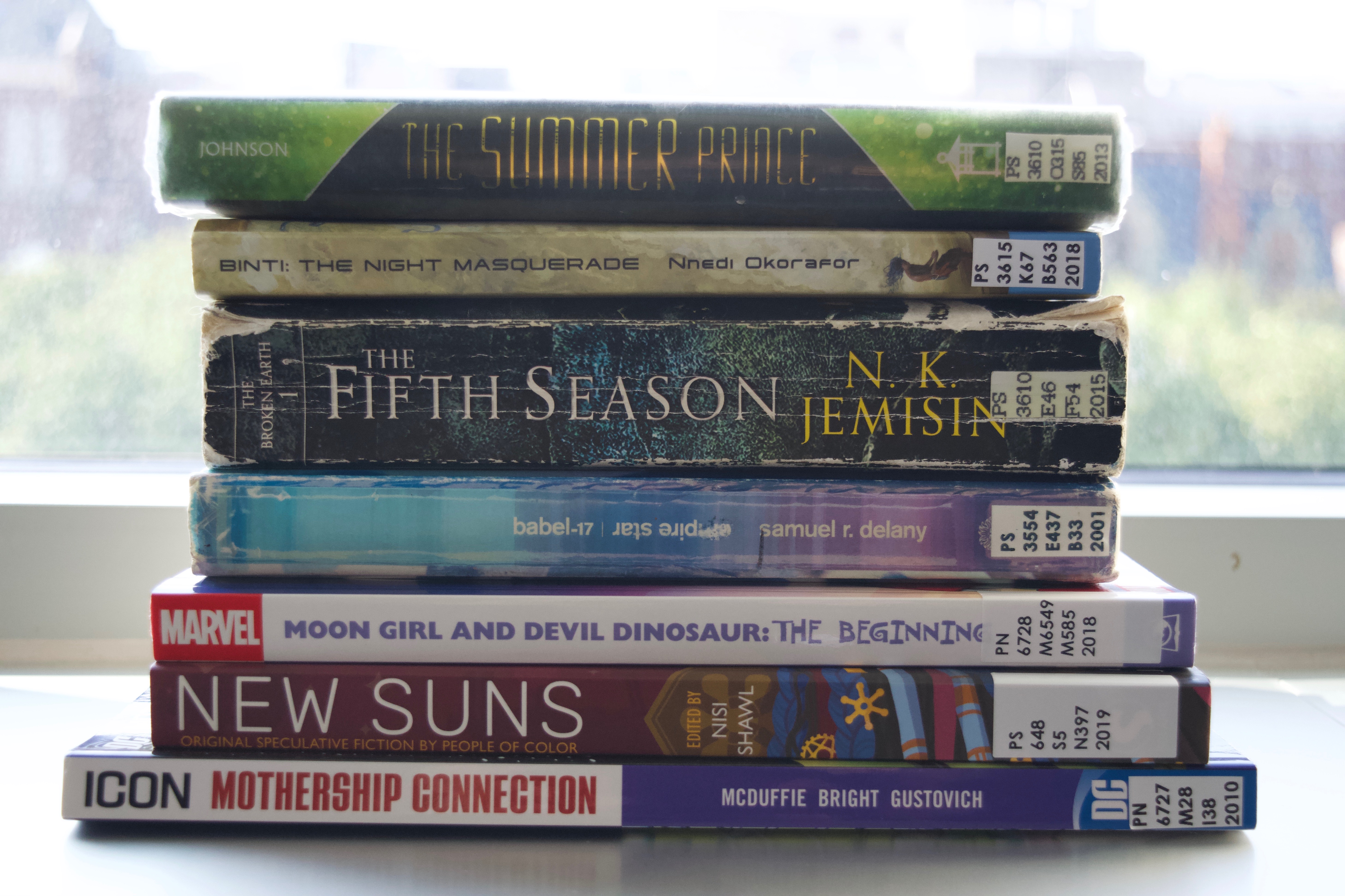 A stack of books with titles including Mothership Connection, New Suns, Moon Girl and Devil Dinosaurs, The Fifth Season, The Night Masquerade, The Summer Prince