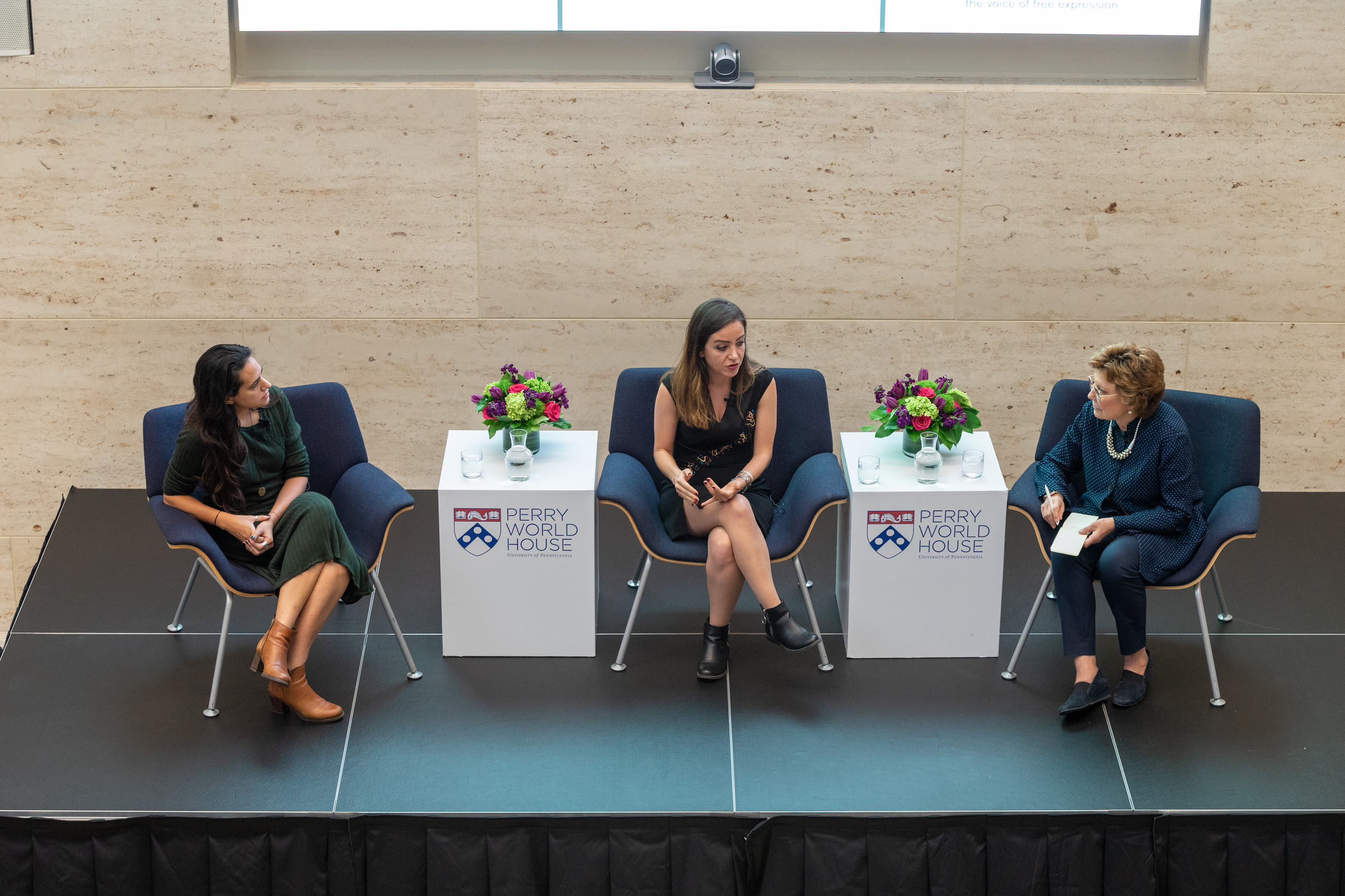 Three people sitting on a stage with endtables between them that read Perry World House. 