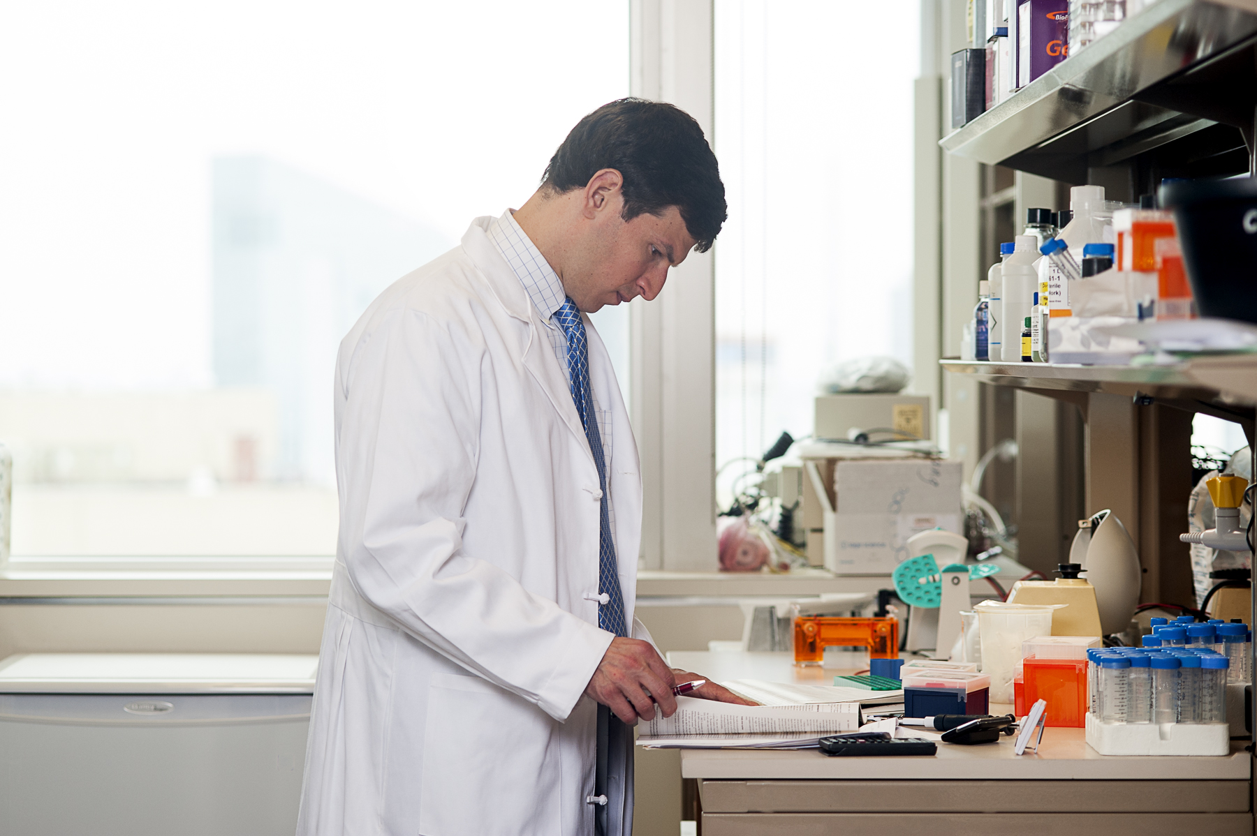 Person in a white lab coat standing at a lab bench looking at a book. Scientific materials are on the table next to and above the person.