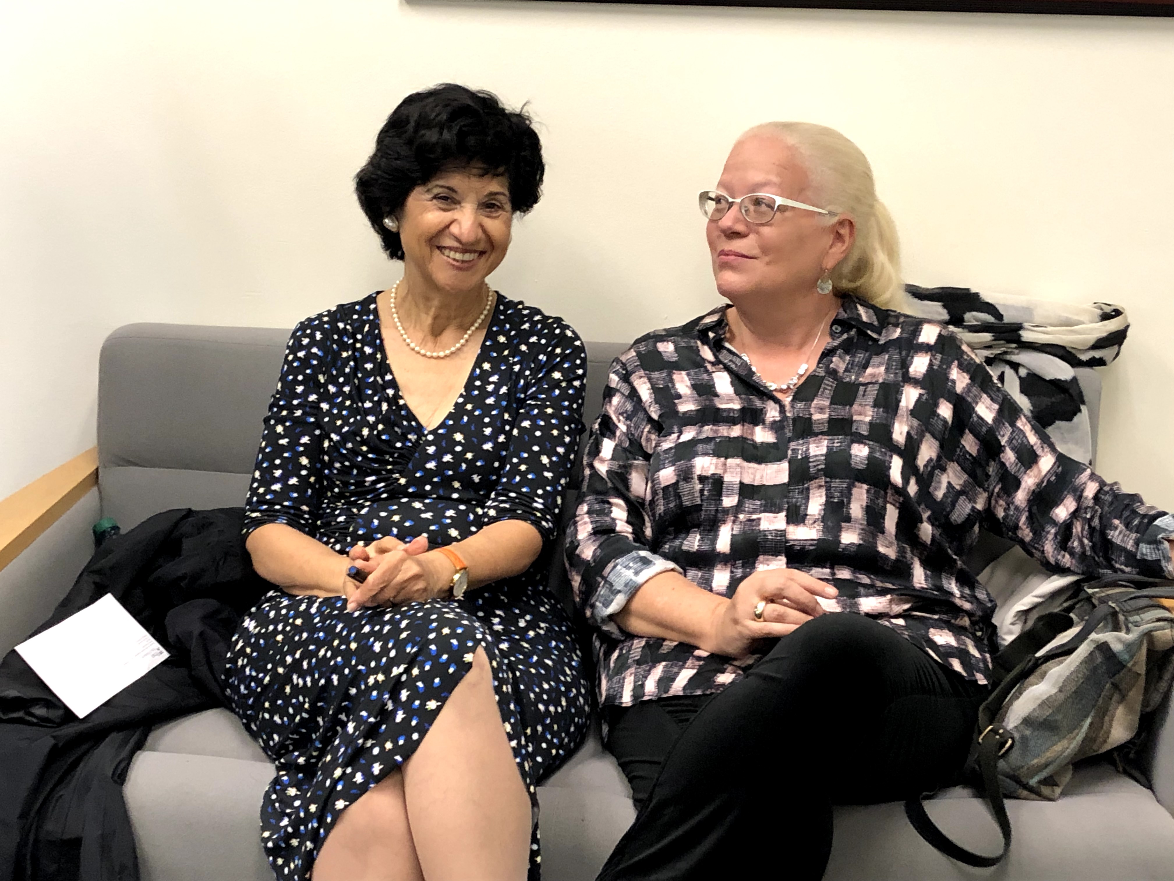 Two people sitting on a gray couch, both with their legs crossed. One faces the camera and smiles, the other looks away at something off camera right.