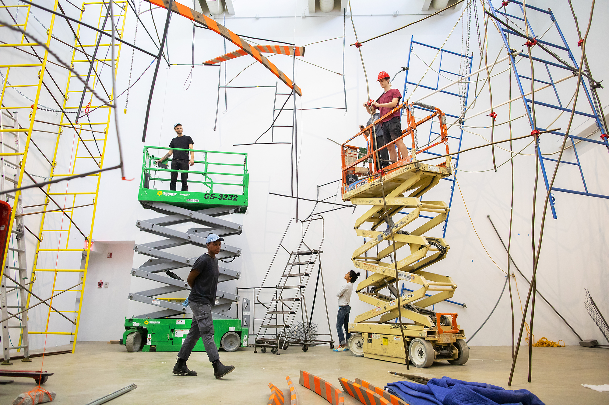 Construction materials hang in an art gallery during installation with two people up on lifts, one person walking, and the artist intently watching. 