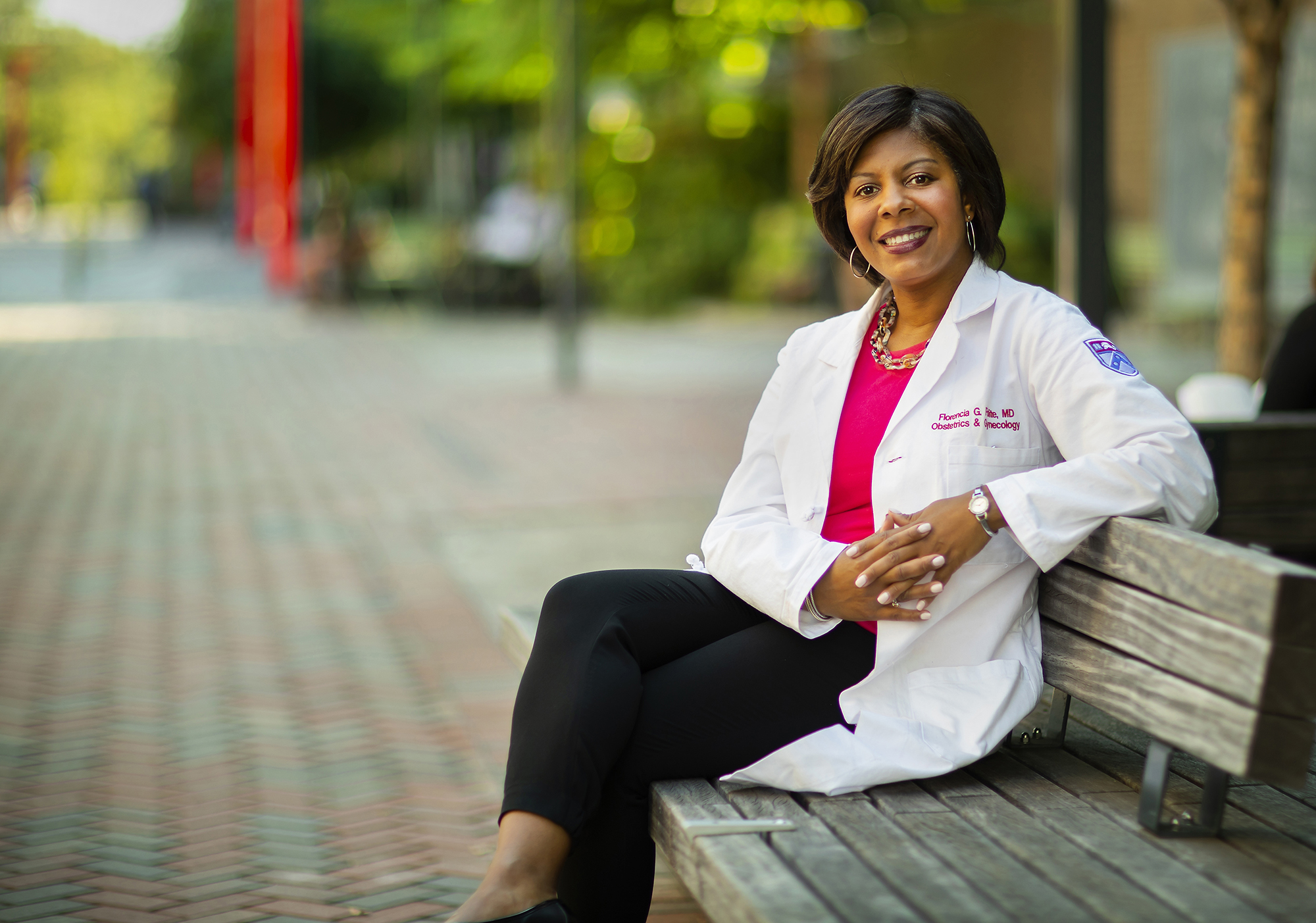 Person in a lab coat sitting on a wooden bench outside. 