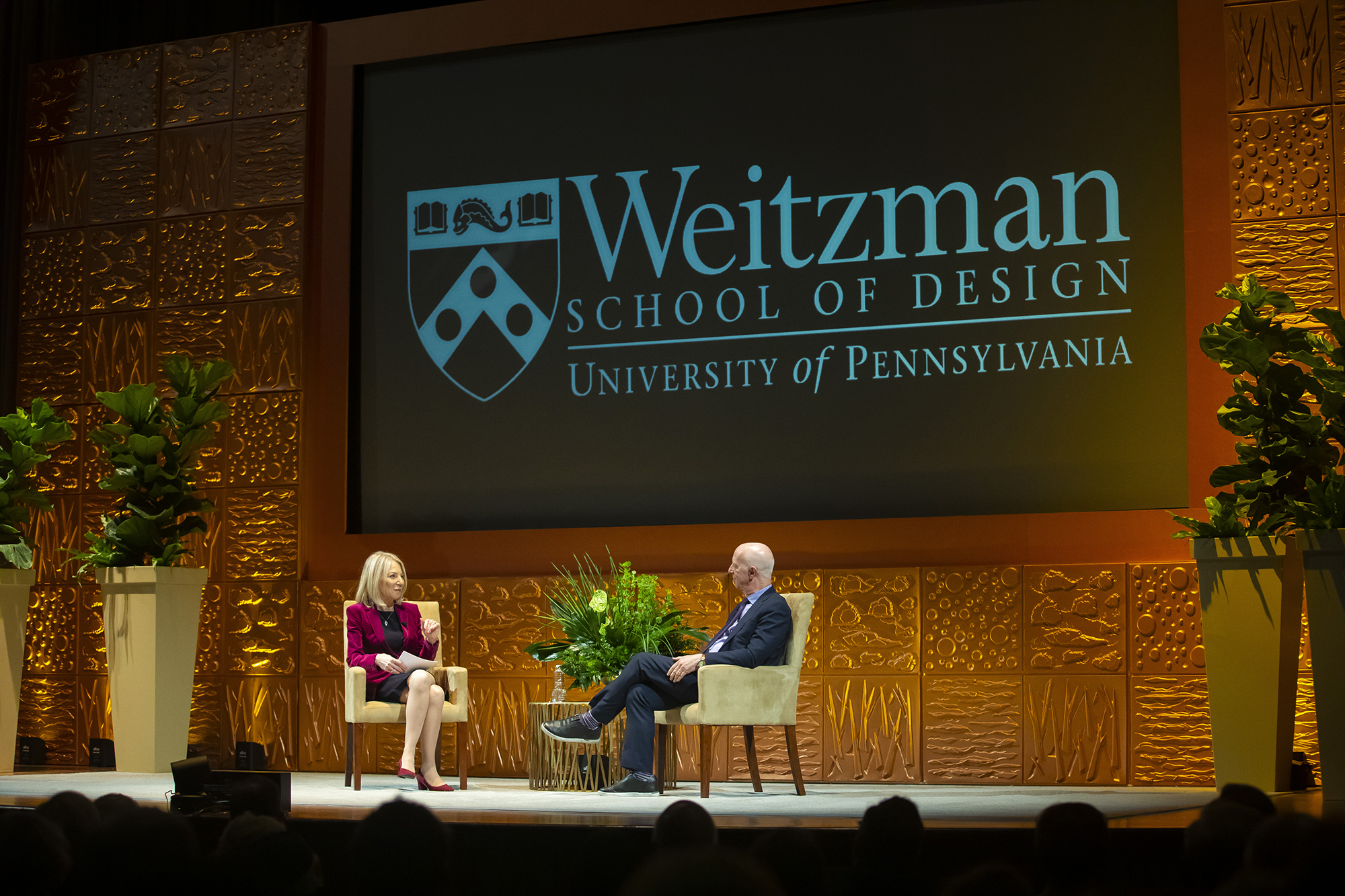 Two people sitting in chairs on a stage with a big sign saying Weitzman School of Design behind them. 