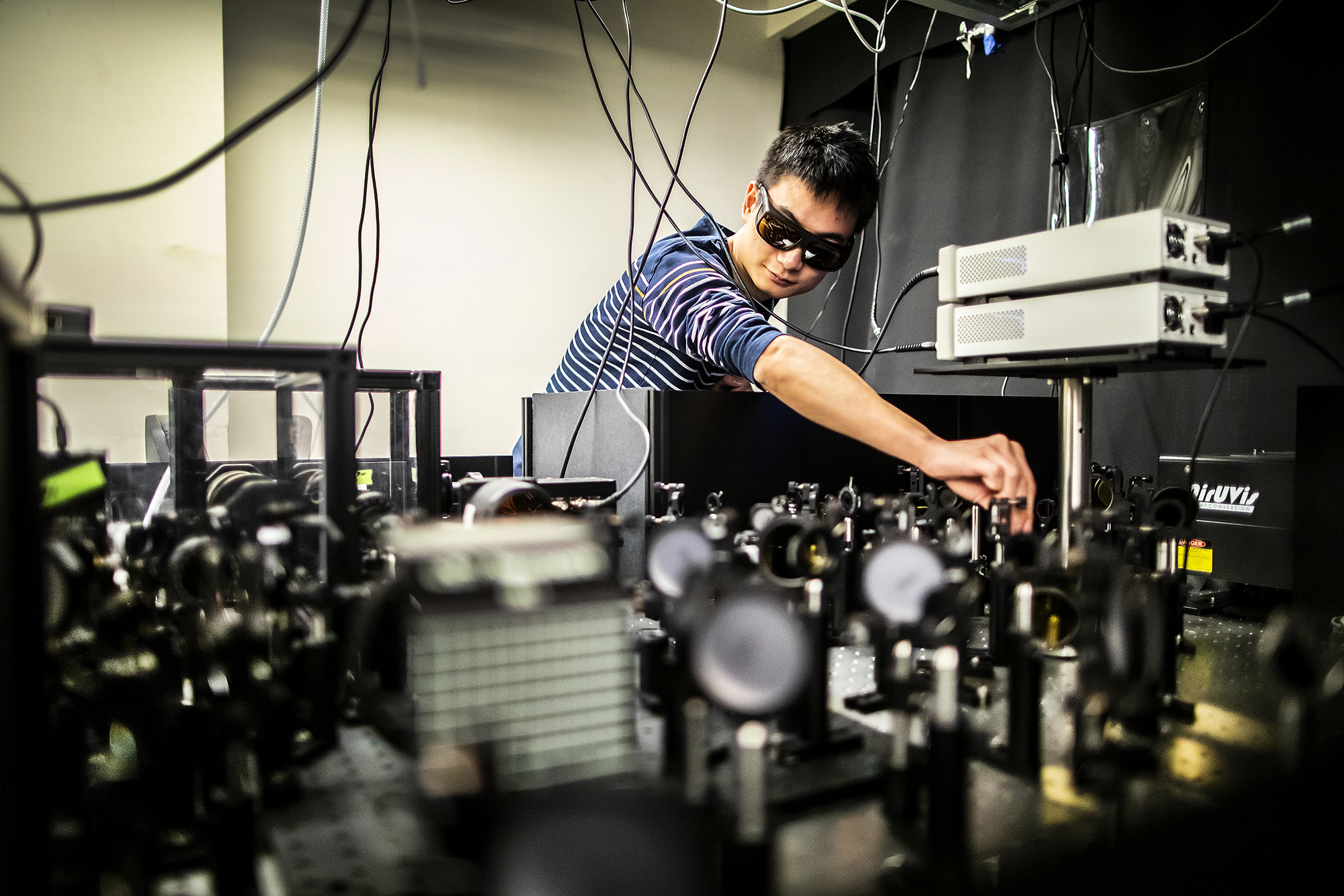 ni adjusts a lens on the top of an optics table covered with lenses and laser boxes