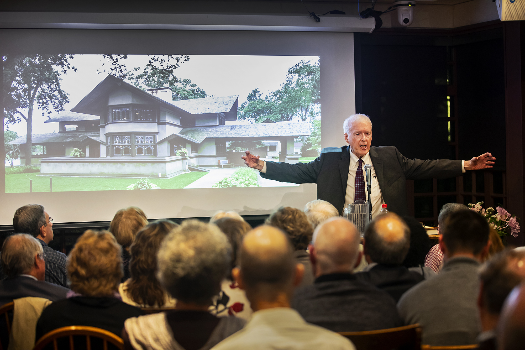 Paul Hendrickson speaking at a podium in front of a seated crowd, his arms outstretched, with a photo of a house on the screen behind him. 