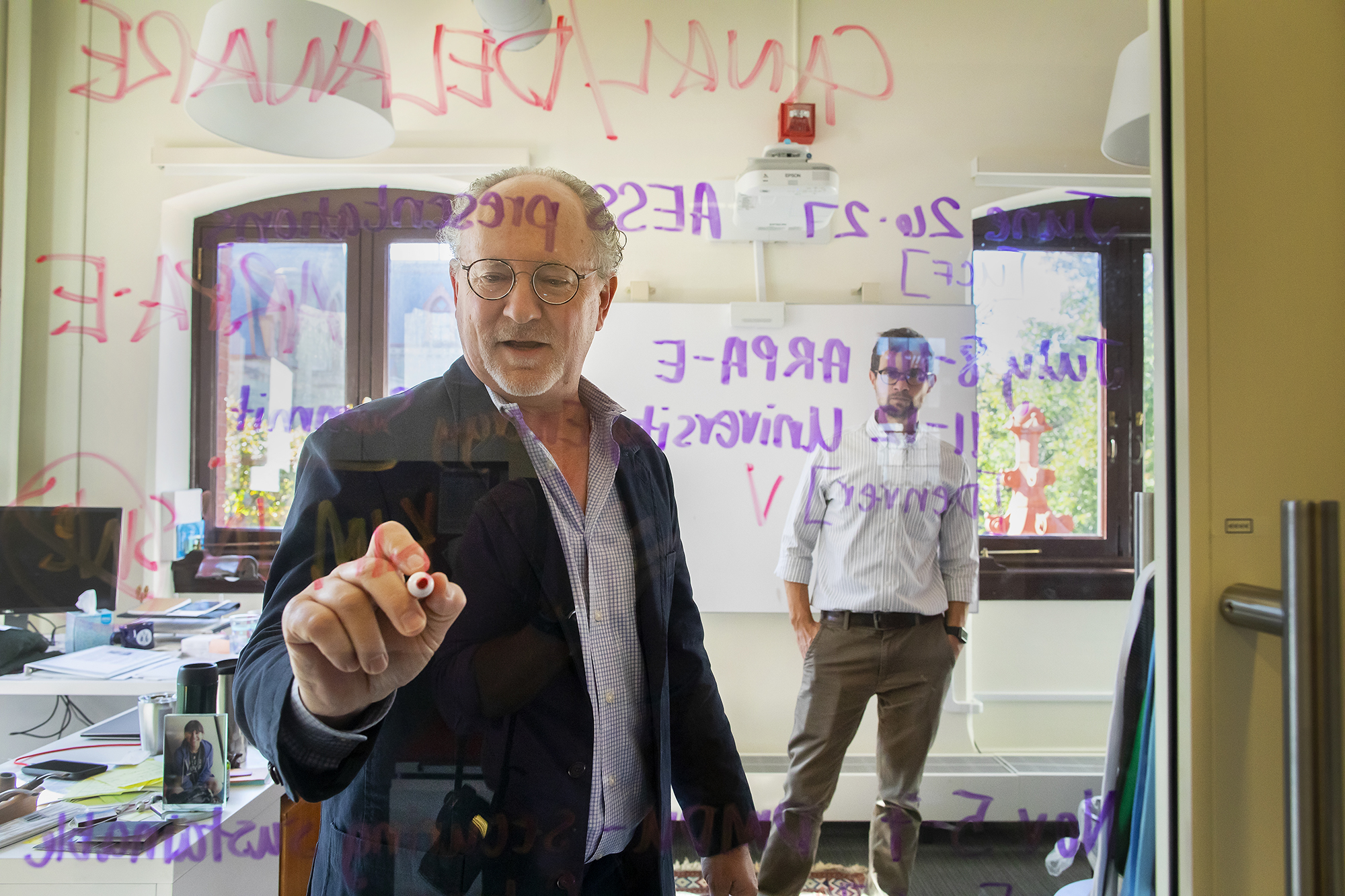 Mark Alan Hughes writes on a clear glass board while a colleague watches in the background.