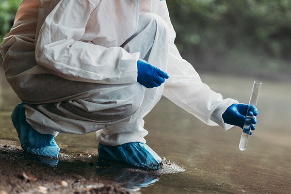 A researcher in protective clothing and gloves gathers a water sample from a river.