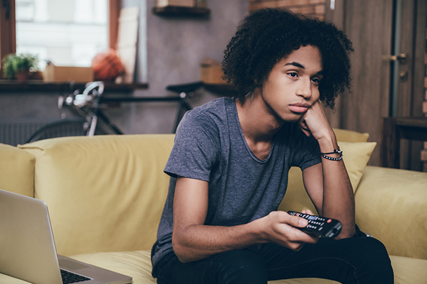 Forlorn teen sits on a couch pointing a television remote control.