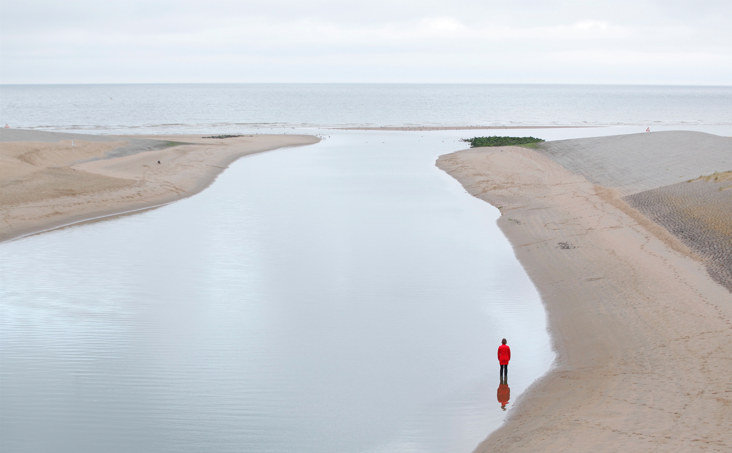 Person in red shirt stands in water looking out at the vast sea